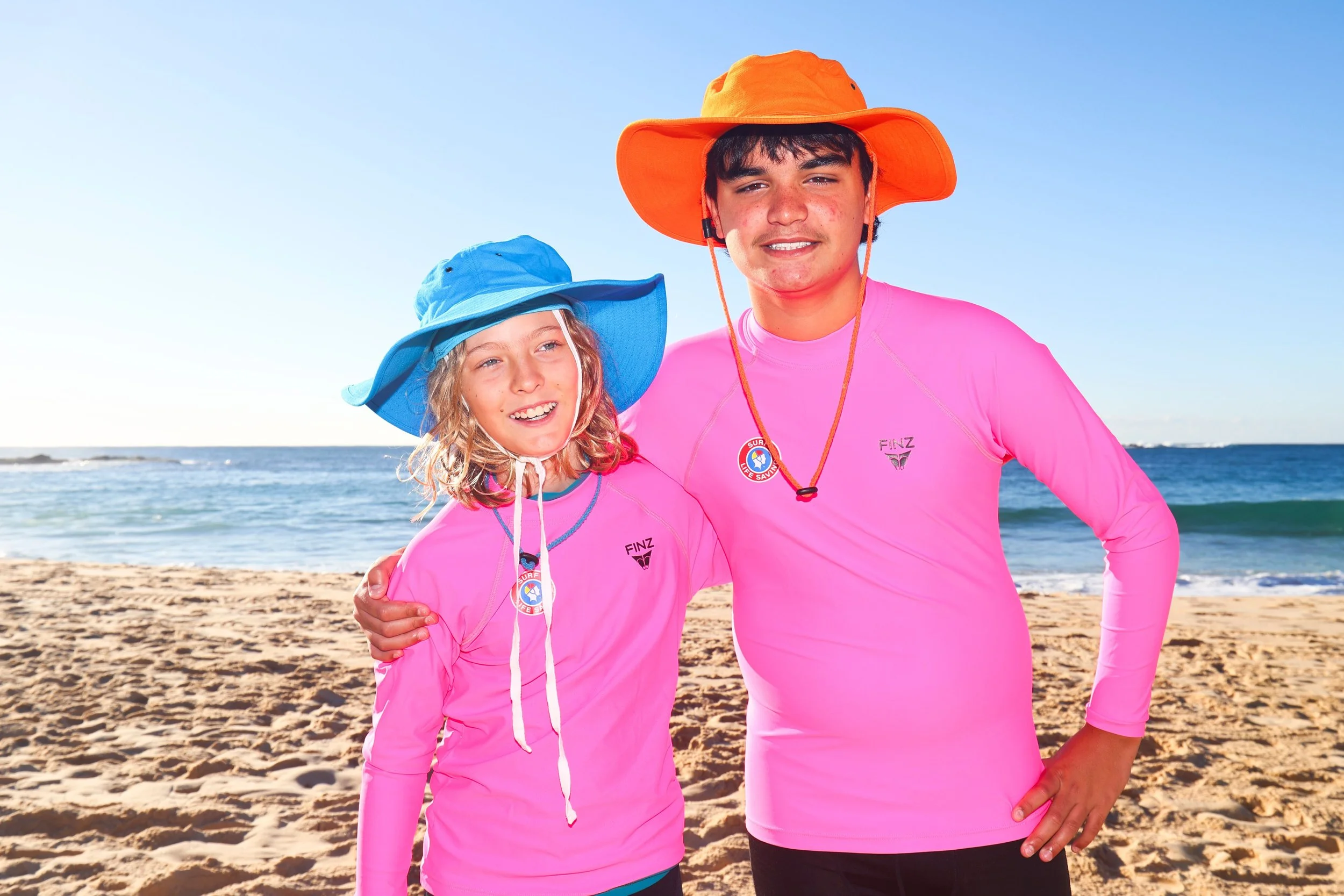 Two young people, a girl and a boy, smiling on a beach, wearing bright pink shirts and wide-brimmed hats for sun protection. The girl has a blue hat and the boy has an orange hat. The ocean and sandy beach are visible in the background.