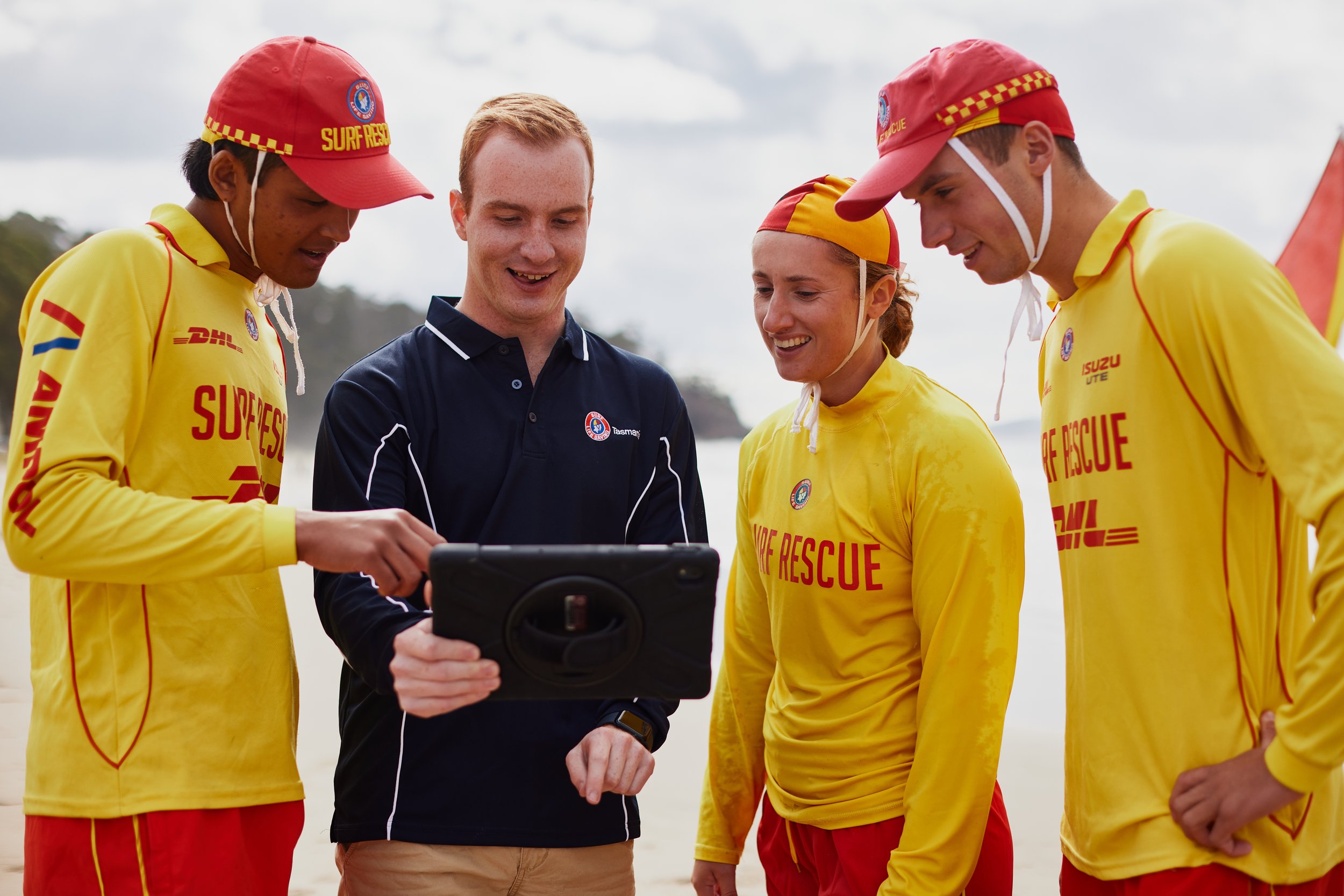 Four rescue workers in yellow uniforms and red hats, along with a man in a navy polo shirt, looking at a device outdoors.