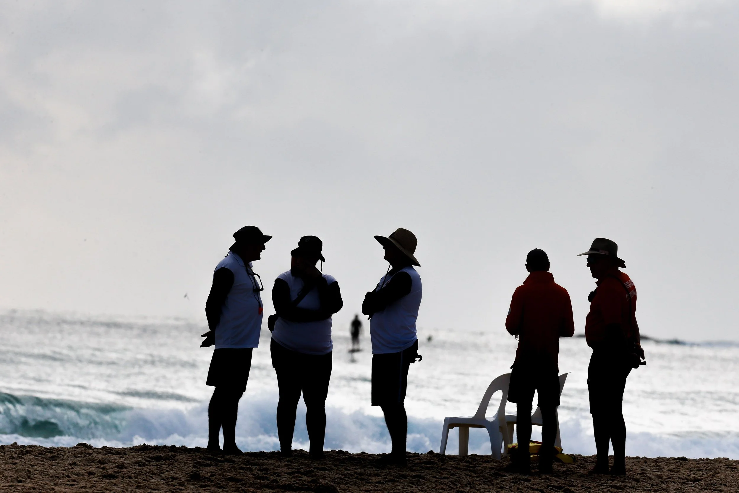 Silhouettes of people standing on the beach near the ocean, some wearing hats, with chairs and surfboards nearby, under an overcast sky.