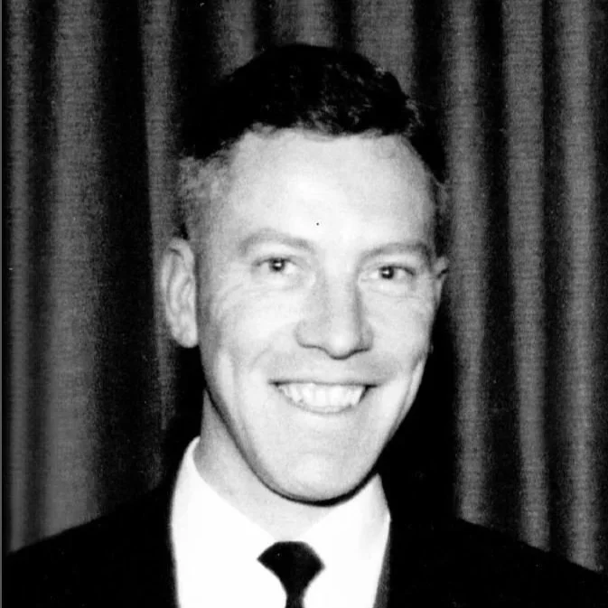 Black and white photo of a smiling man in a suit and tie standing in front of curtains.