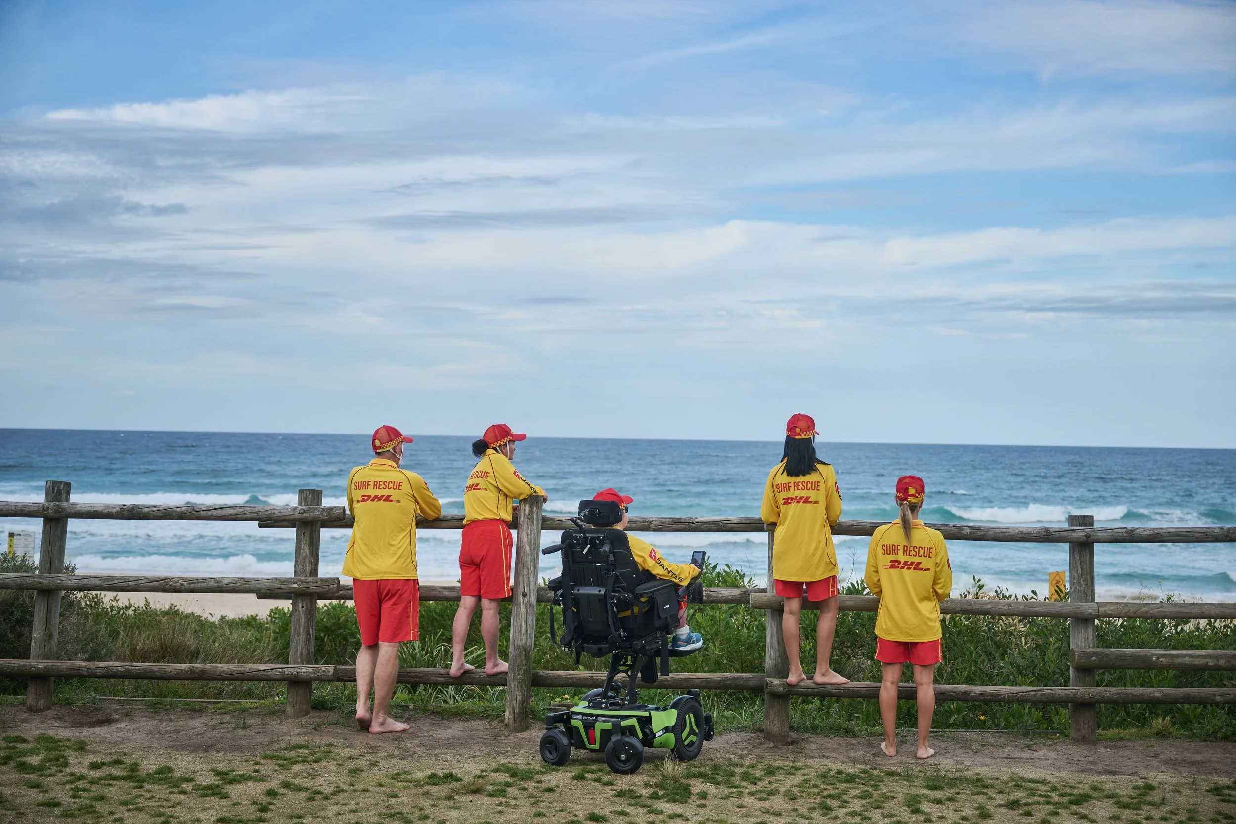 Surfers and rescue team members in yellow and red uniforms standing by a wooden fence, looking at the ocean on a cloudy day with a few rescue vehicles nearby.