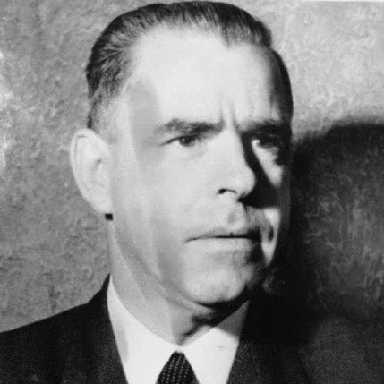 Black and white portrait of a man with slicked-back hair, wearing a suit and tie, looking to his left with a serious expression.