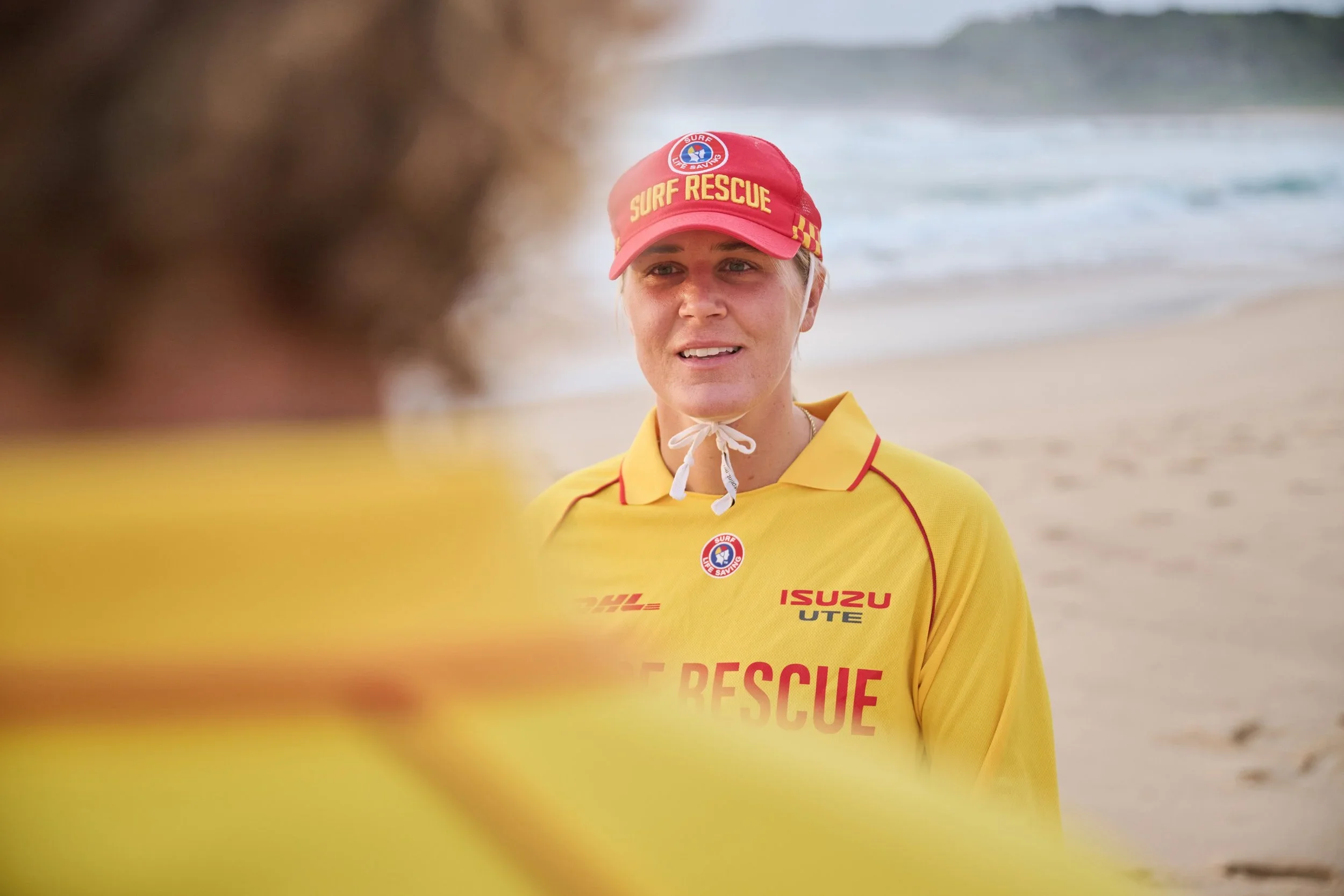 A female surf rescue worker in a yellow uniform and red cap with 'Surf Rescue' written on it, standing on a beach with ocean waves in the background.