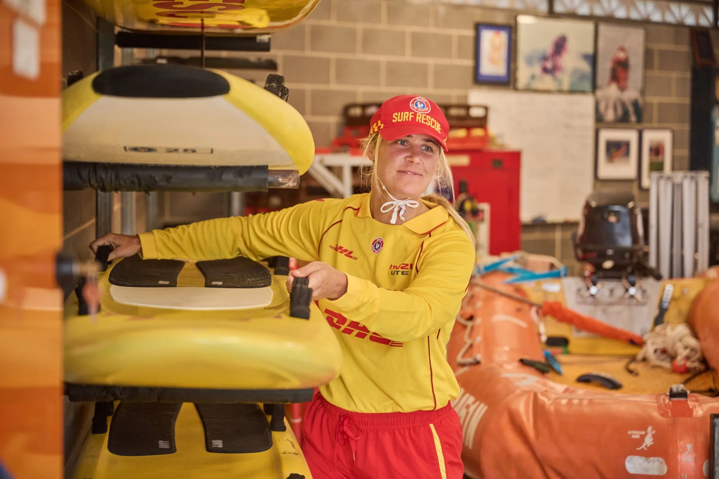 A woman in a yellow surf rescue shirt and red cap with matching red shorts stands next to a yellow surfboard inside a storage room. The background shows various surf rescue equipment.