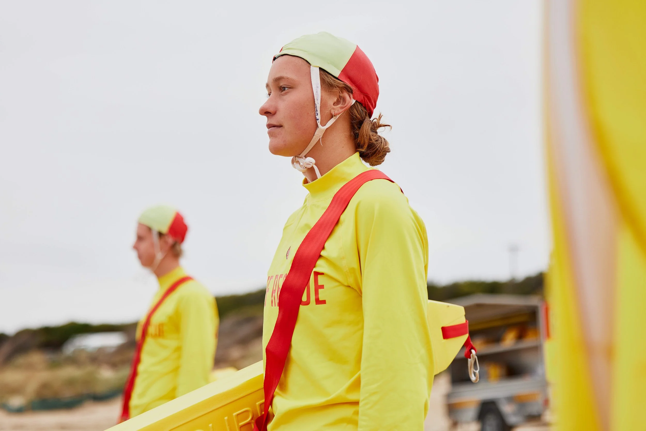A woman in a yellow jacket and red life vest wearing a red and green helmet at a beach location. She is standing in profile with a focused expression.