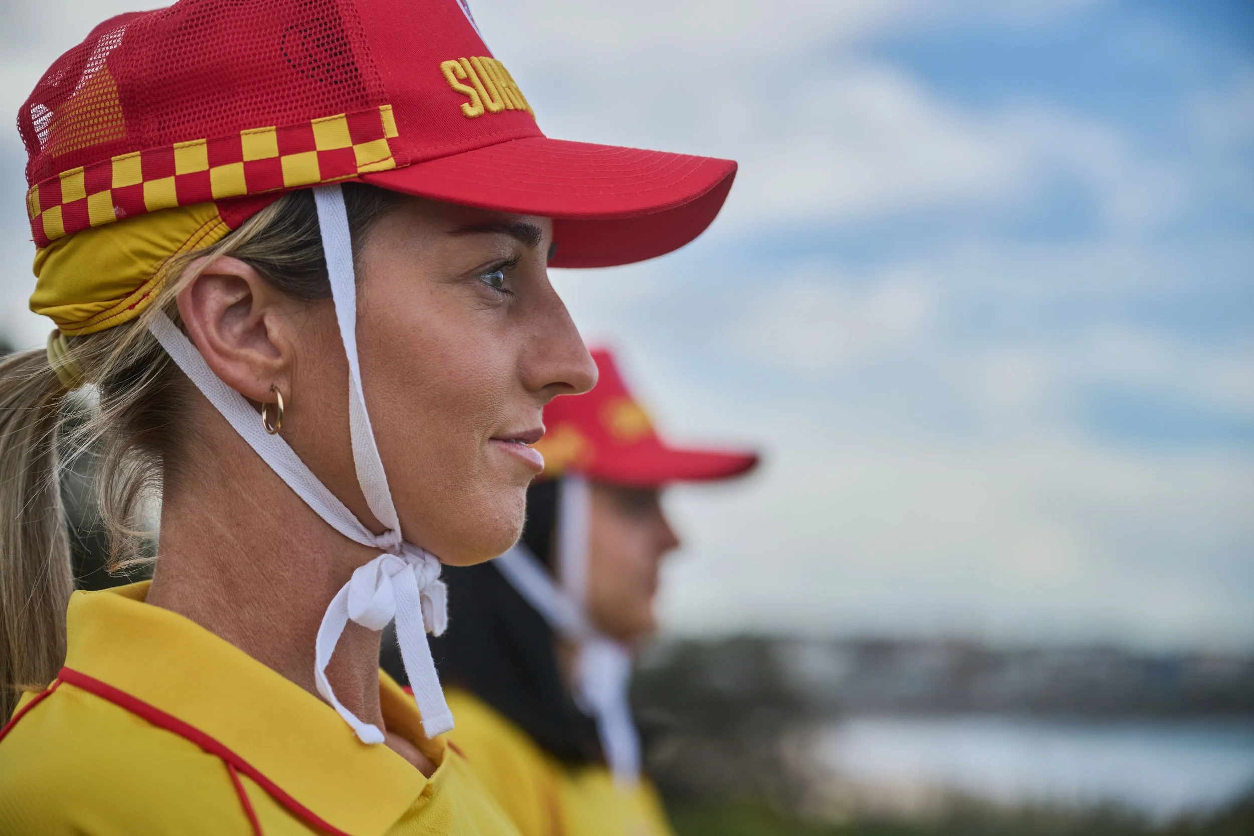 A woman in a yellow and red uniform with a red cap that has yellow checkered pattern and the word 'Surf' on it, standing outdoors with a blue sky in the background.