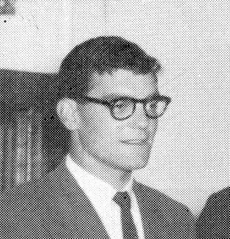 Black-and-white photo of a man wearing glasses, a suit, and a tie, sitting in a chair.