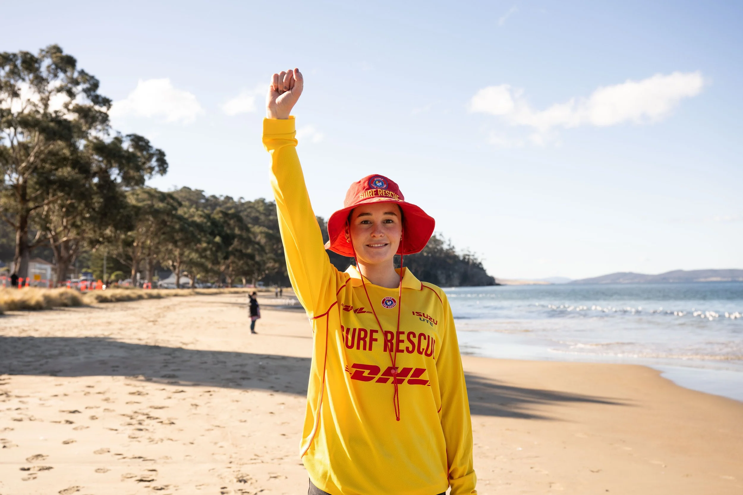 A young woman in a yellow surf rescue uniform and red sun hat raises her hand in the air on a sandy beach with trees, water, and a cloudy sky in the background.