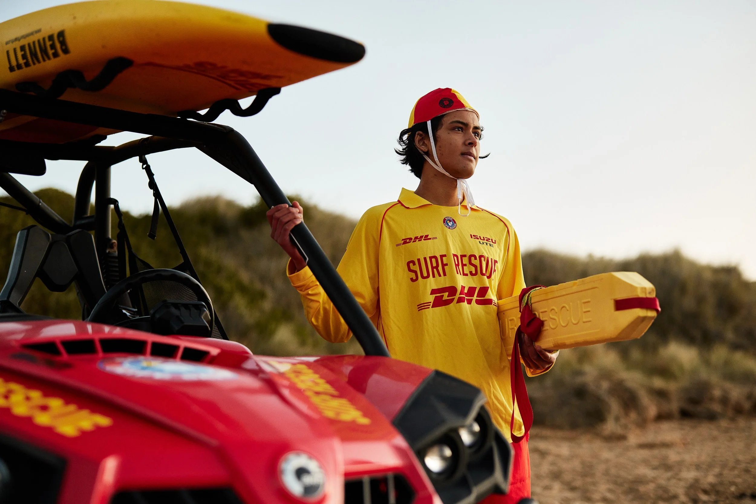 A young man dressed in a yellow surf rescue uniform with red text and logos, including DHL and Isuzu, holds a yellow rescue kit and stands outdoors near a red rescue ATV. He is wearing a red and yellow helmet and looks into the distance.