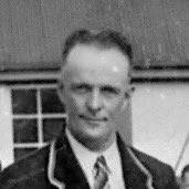 Black and white photo of a man in a suit standing outdoors near a house with a window.
