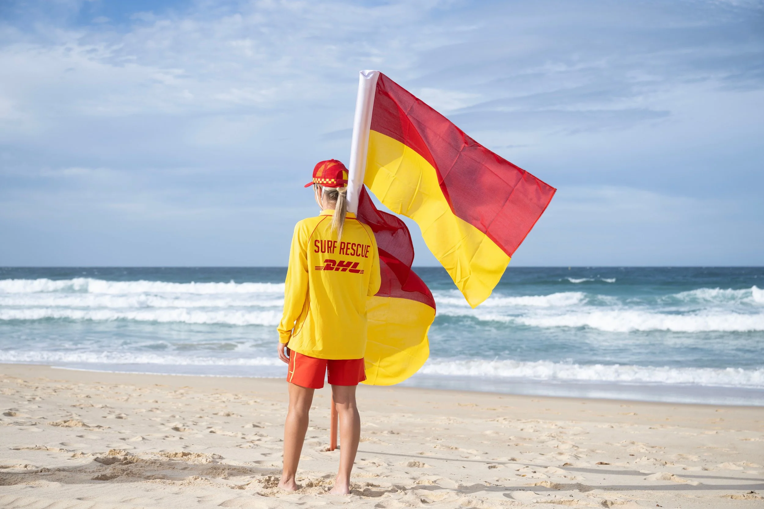 A person in a yellow surf rescue jacket and red shorts standing on a sandy beach, holding a red and yellow flag, with the ocean in the background.
