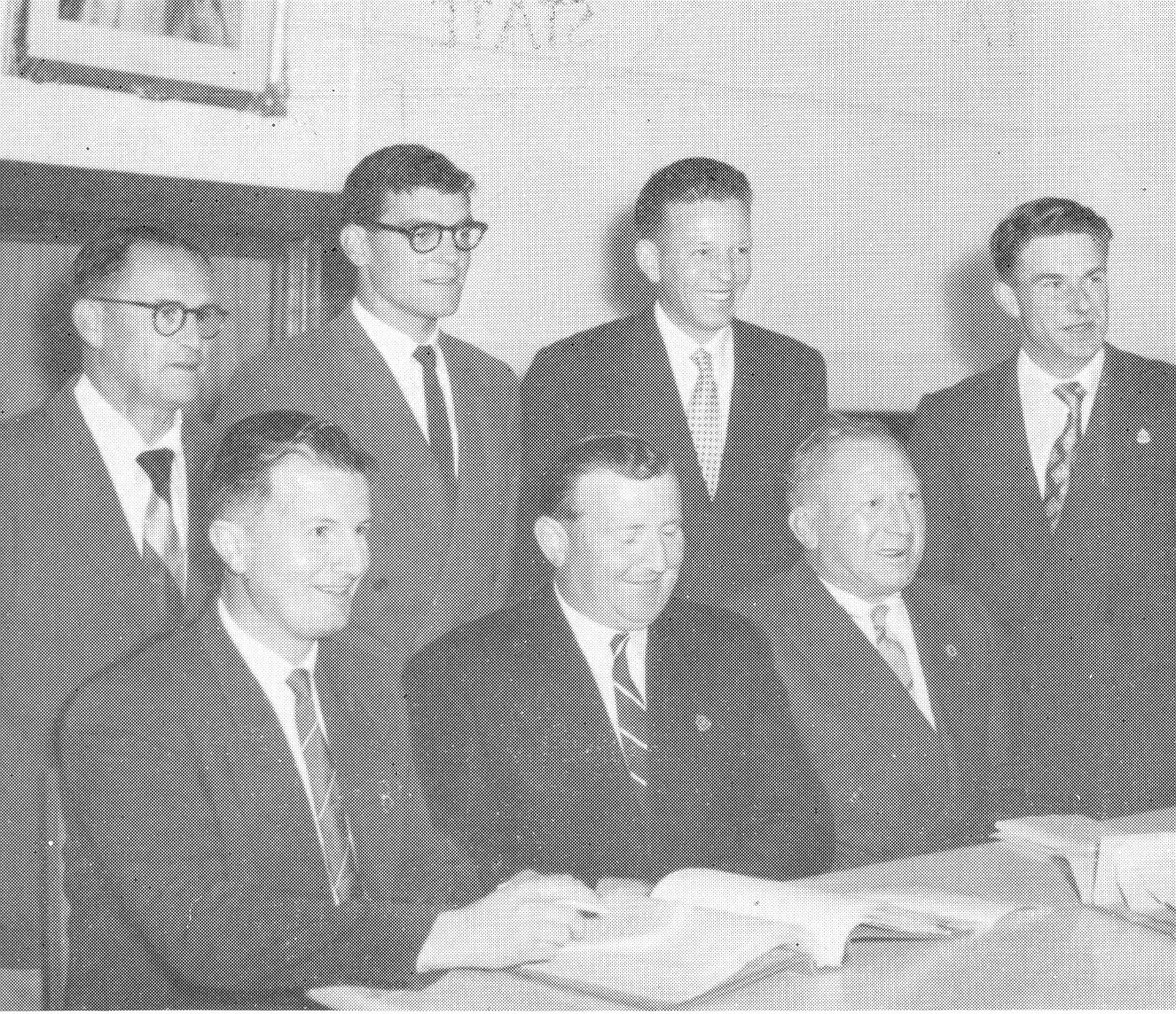 Black and white photo of seven young men in suits and ties sitting and standing around a desk, smiling at the camera.