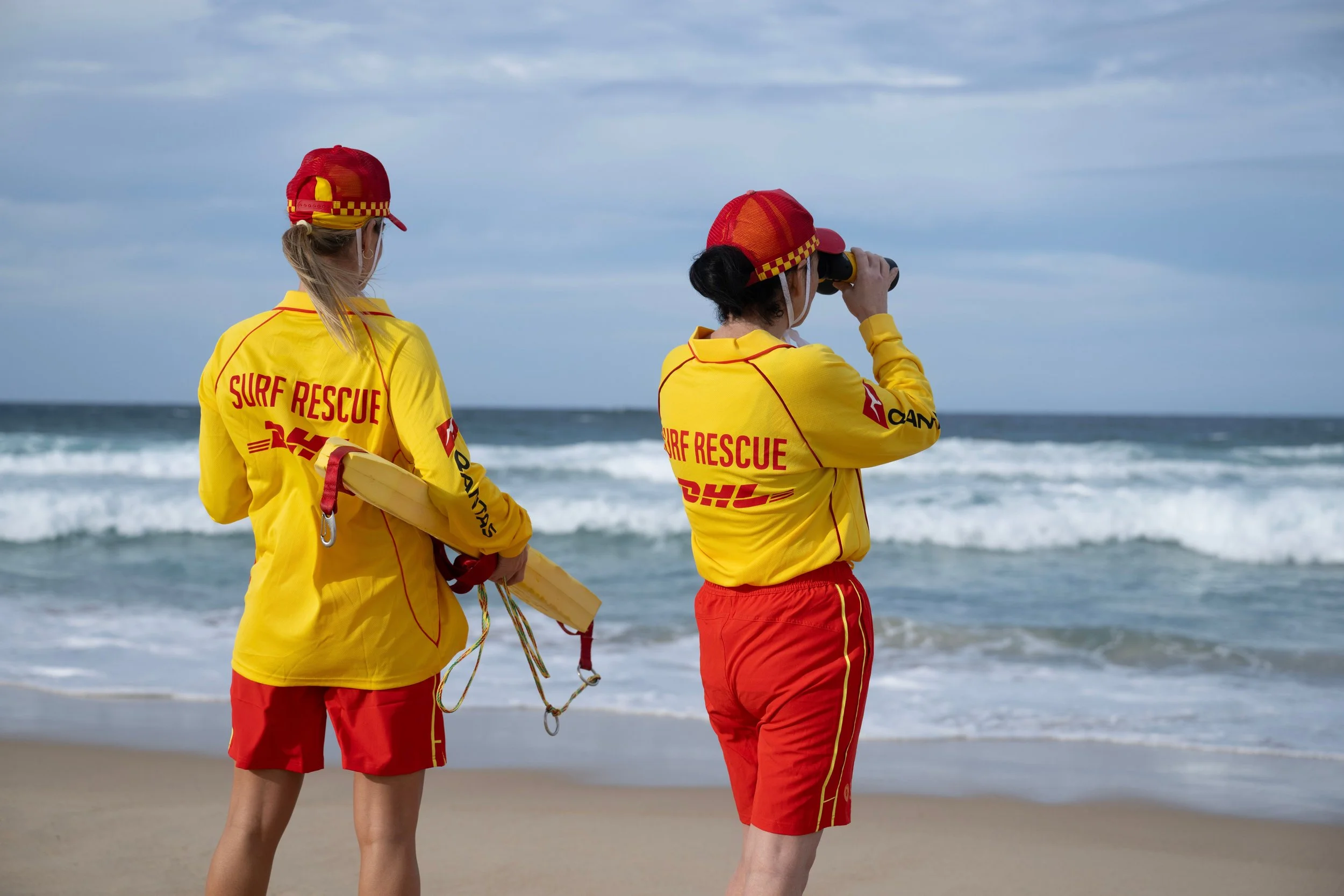 Two surf rescue team members in yellow jackets with red and yellow checkered hats, standing on a beach, one looking through binoculars, with ocean waves and a cloudy sky in the background.