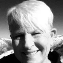 A black and white close-up photo of a smiling person with short hair, outdoors with mountains in the background.