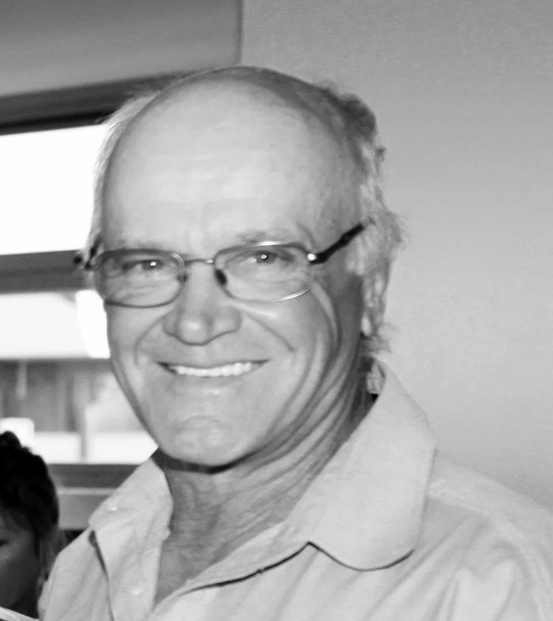 A smiling older man with glasses, wearing a light-colored collared shirt, standing indoors near a window.