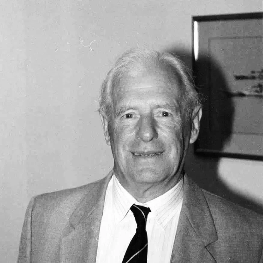 Black and white photo of an older man with light-colored hair, dressed in a suit and tie, standing indoors in front of a plain wall and a framed picture.