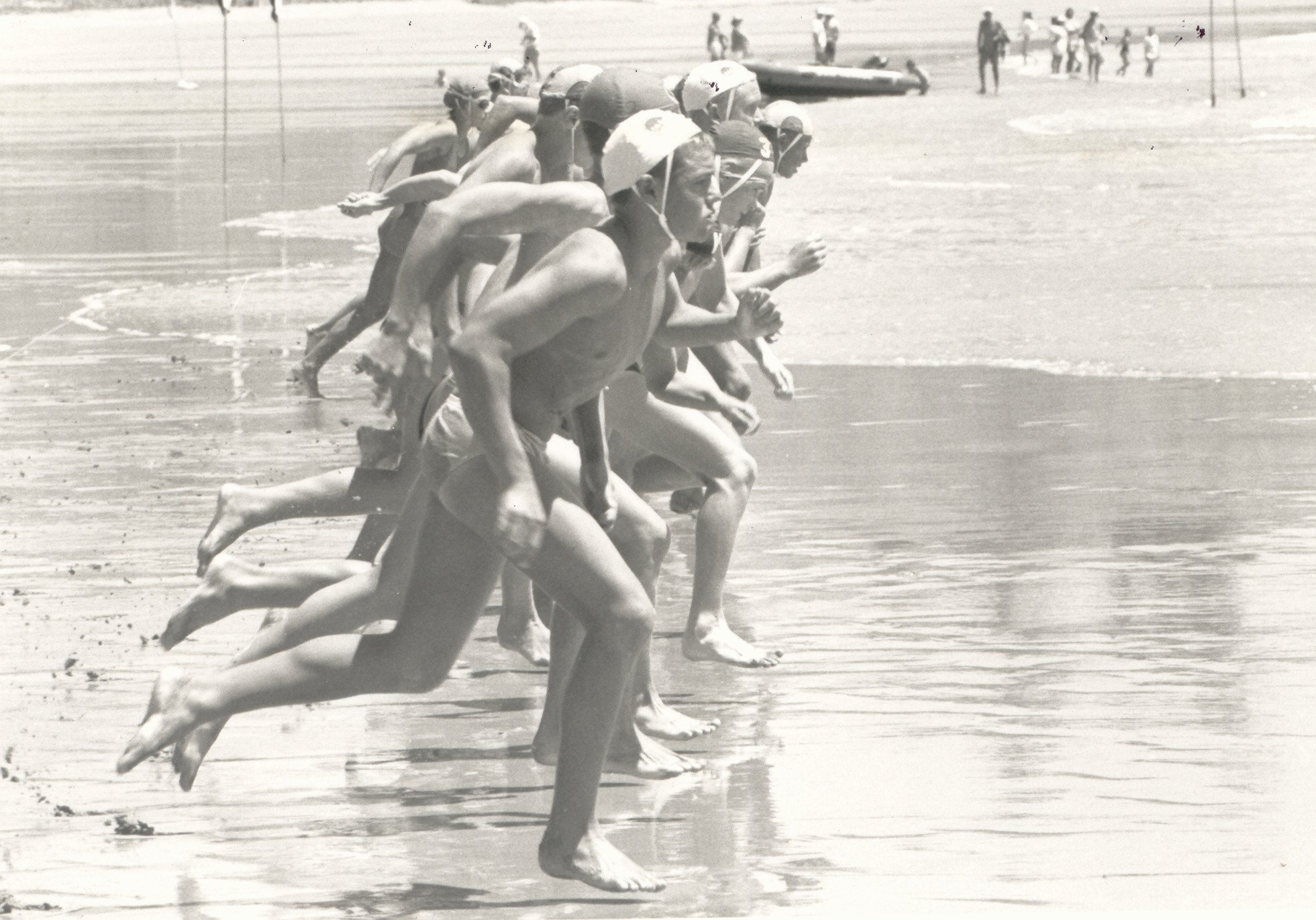 A black and white photograph of a group of people, likely male athletes, running on a beach in a race. They are wearing swim caps, some with goggles, and are tanned, shirtless, in swimwear. The scene captures the moment as they are in motion along the shoreline, with the ocean and distant figures in the background.