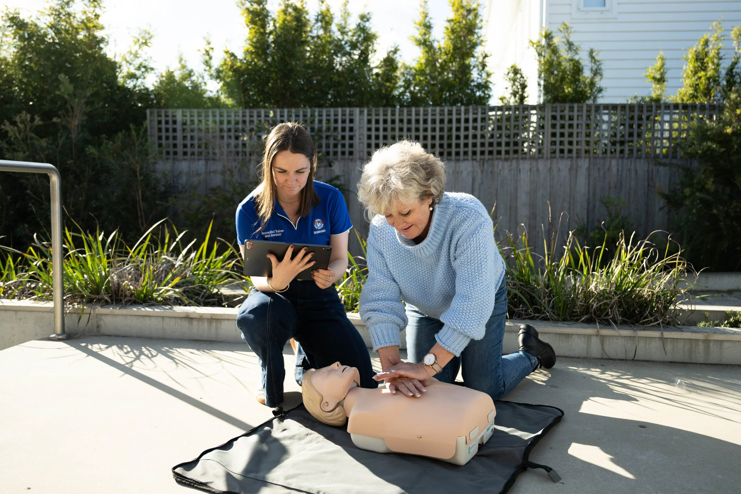 Two women practicing CPR on a training mannequin outdoors during daylight.