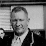 Black-and-white photo of a man with short hair, wearing a collared shirt and blazer, standing outdoors with a blurred background.