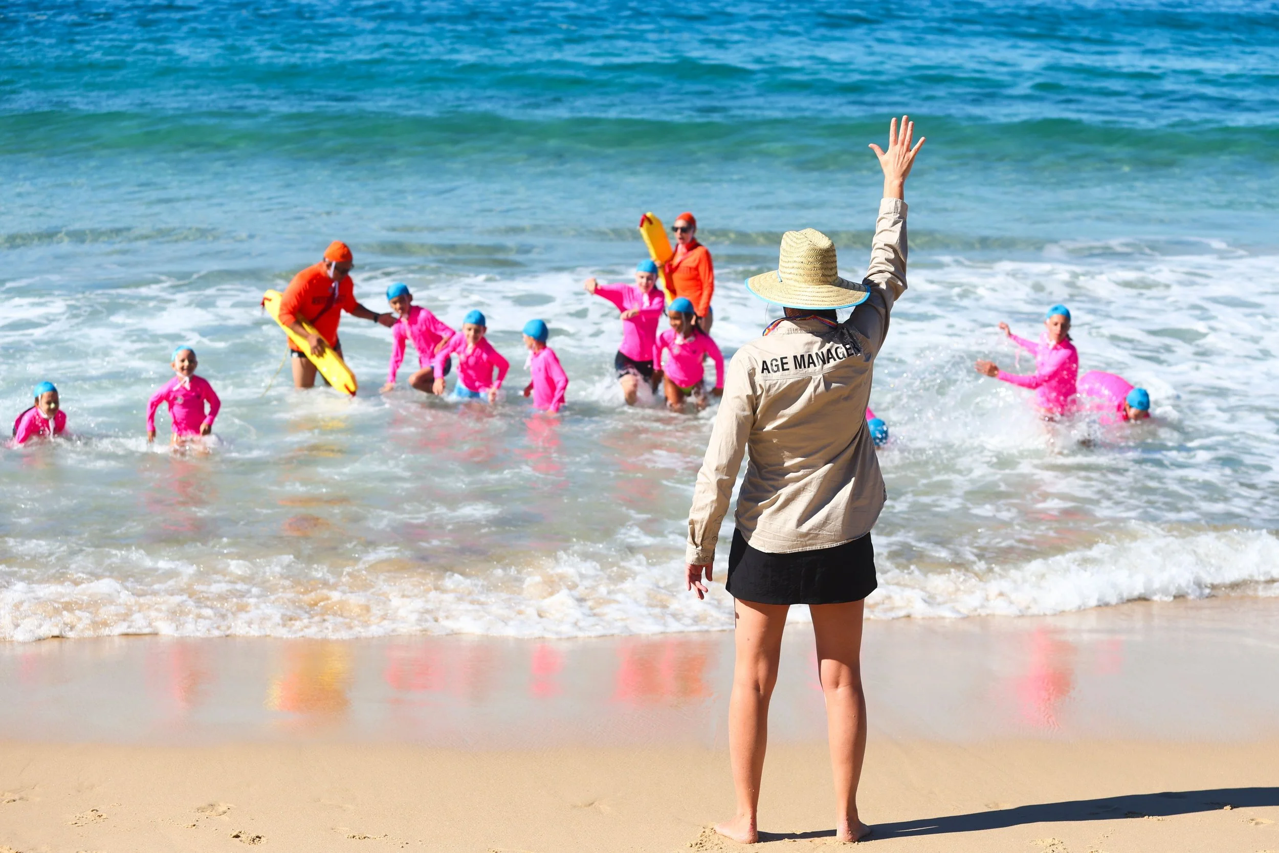 A woman wearing a wide-brimmed hat with a neck cover, a beige jacket labeled 'AGE MANAGER,' and black shorts stands on the beach, raising her arm, while a group of children in pink wetsuits and blue swim caps enjoy the shallow ocean waves, supervised by two adults.