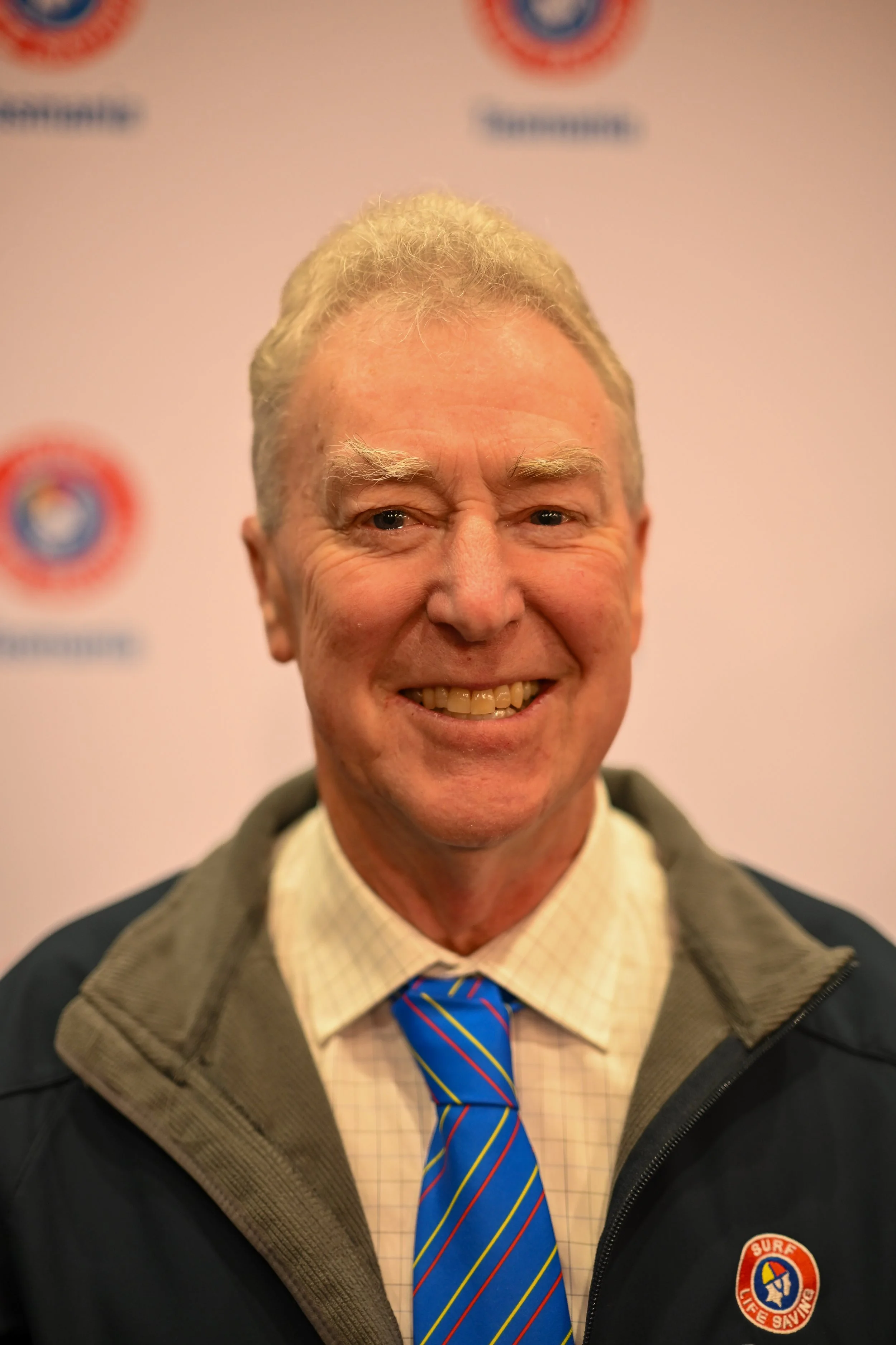 A smiling man with gray hair, wearing a beige checked shirt, a blue tie with yellow and red stripes, and a brown and black jacket with a surfing life-saving badge on it. The background features a blurred banner with logos and text.