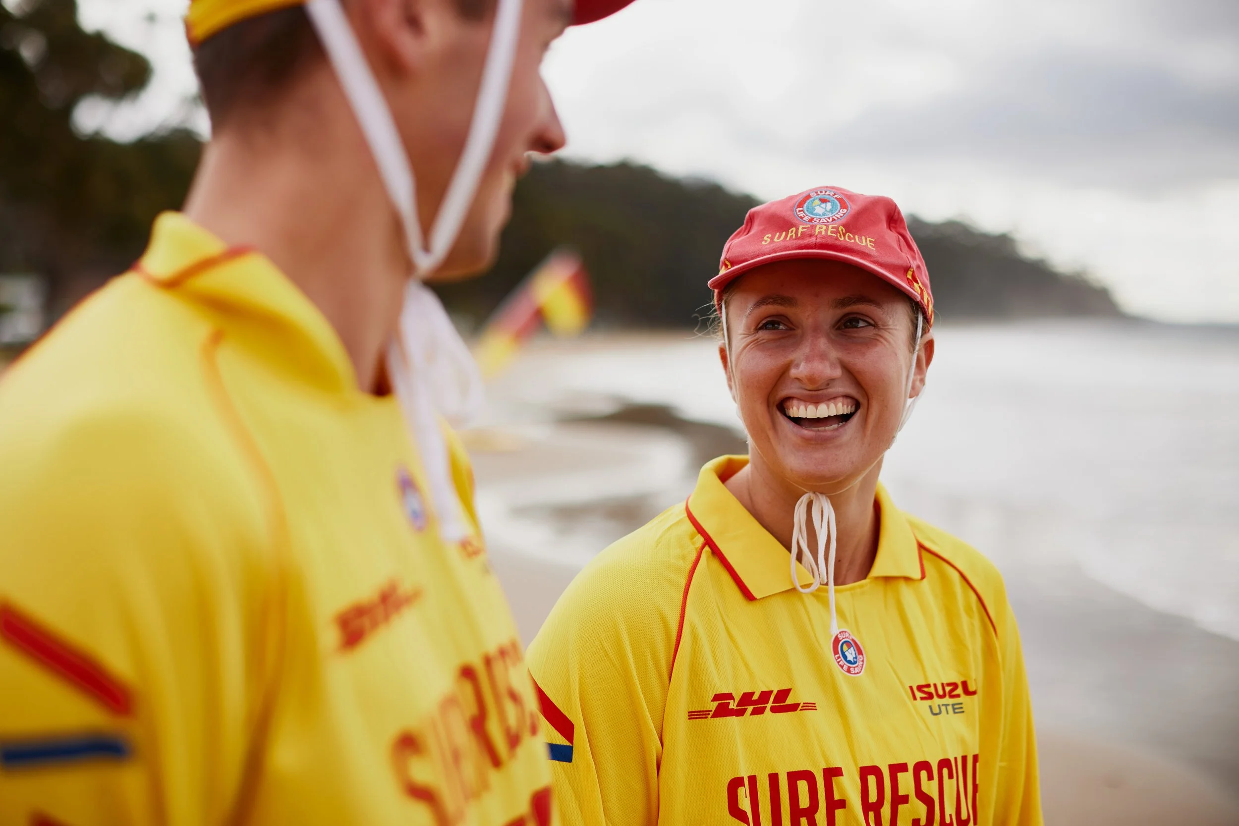 Two women in yellow surf rescue uniforms smiling and talking on a beach, with cloudy sky and water in the background.