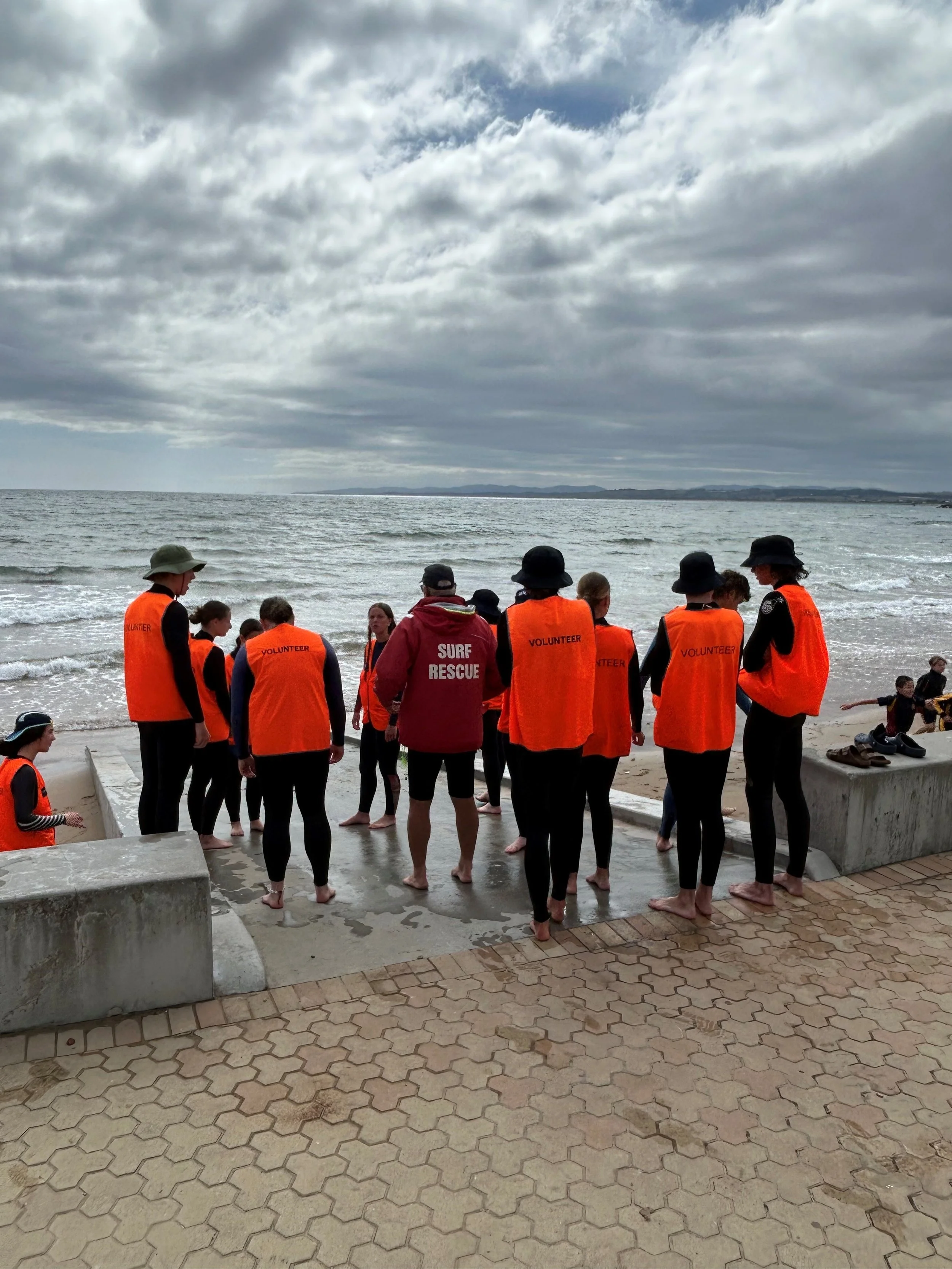 Group of people in orange life jackets and wetsuits standing on a beach near the ocean, with cloudy skies overhead.