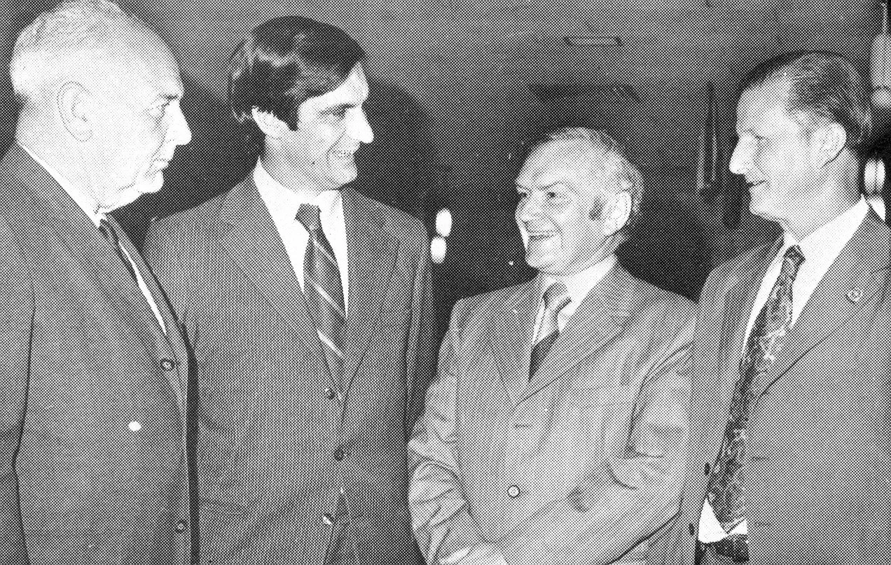 Four men in suits engaged in conversation indoors, with a dark background.