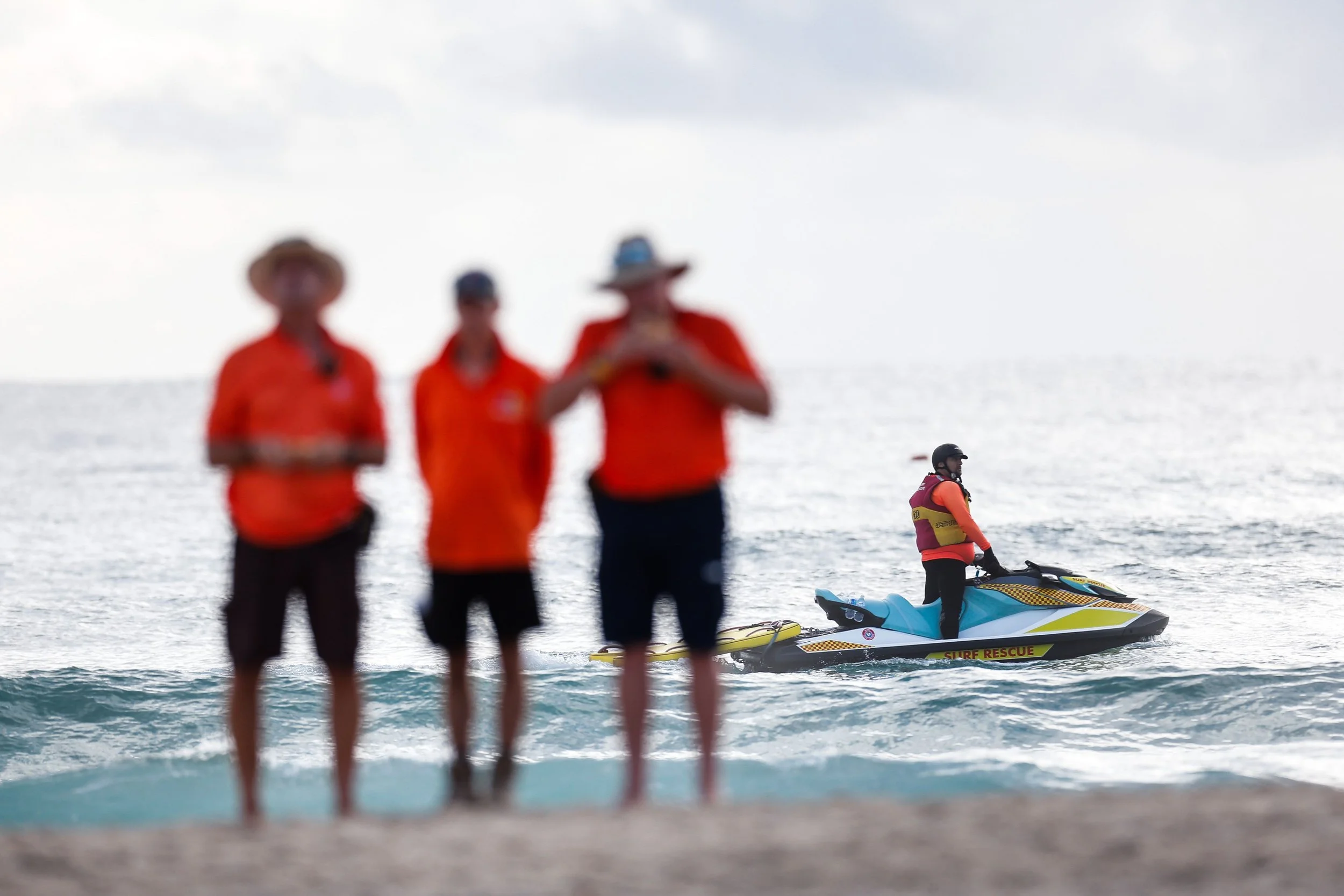 People at the beach, with three individuals in the foreground blurred, wearing orange shirts, and two people in the background near the water, one on a jet ski labeled 'Surf Rescue' and the other steering a boat, all under a cloudy sky.