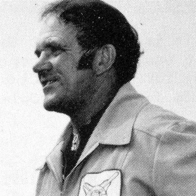 Black and white photo of a man in a jacket with a patch on the sleeve, smiling and looking to the side.