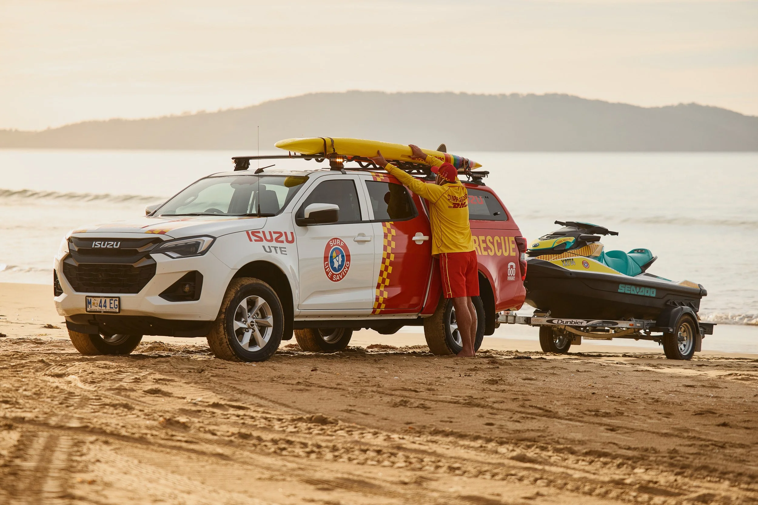 A rescue worker in yellow uniform loading a yellow surfboard onto a vehicle at the beach, with a jet ski on a trailer attached to the vehicle and calm water in the background during sunset.