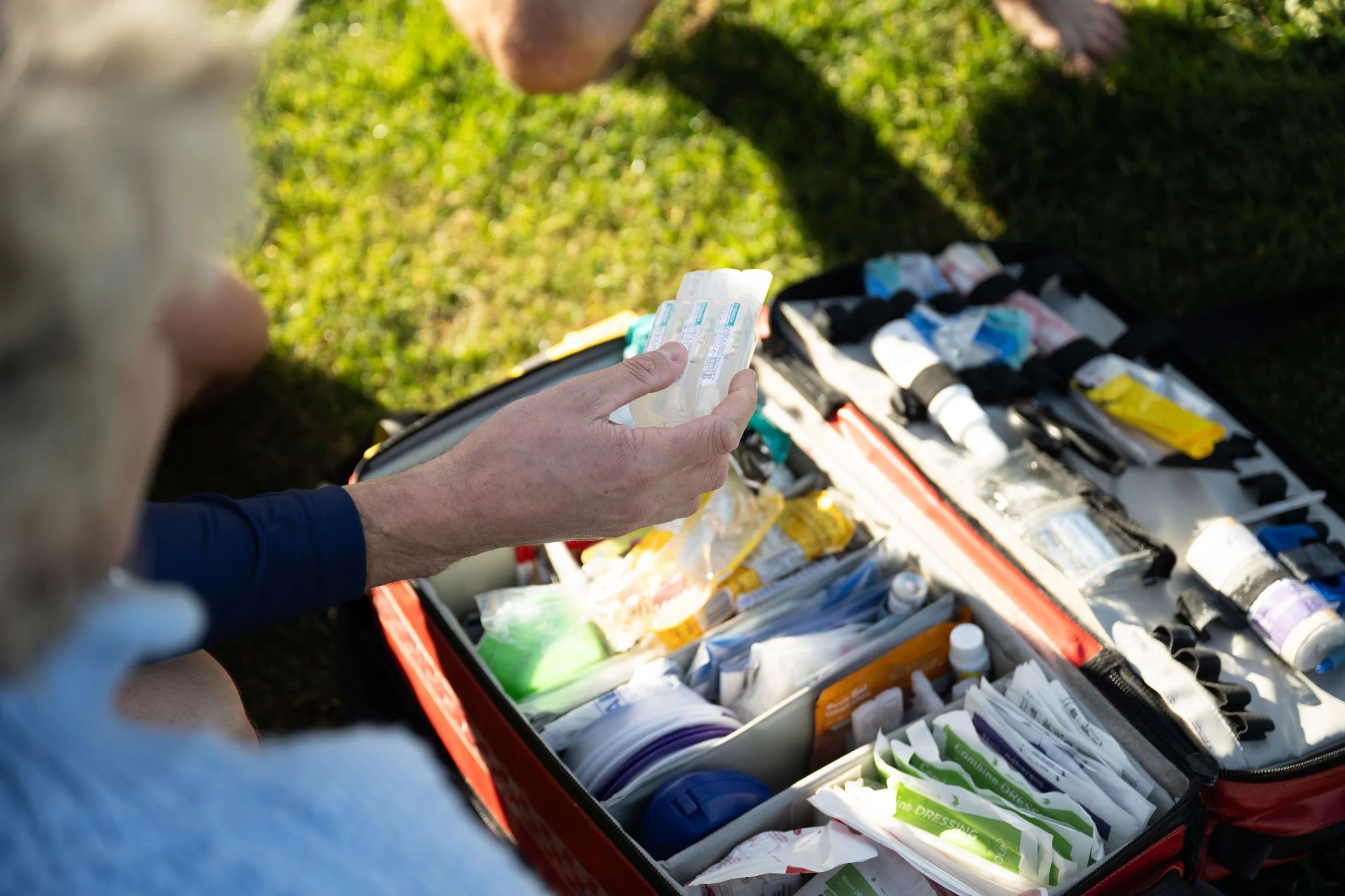 Person holding a pack of medical syringes and a first aid kit opened on grass, with medical supplies inside.