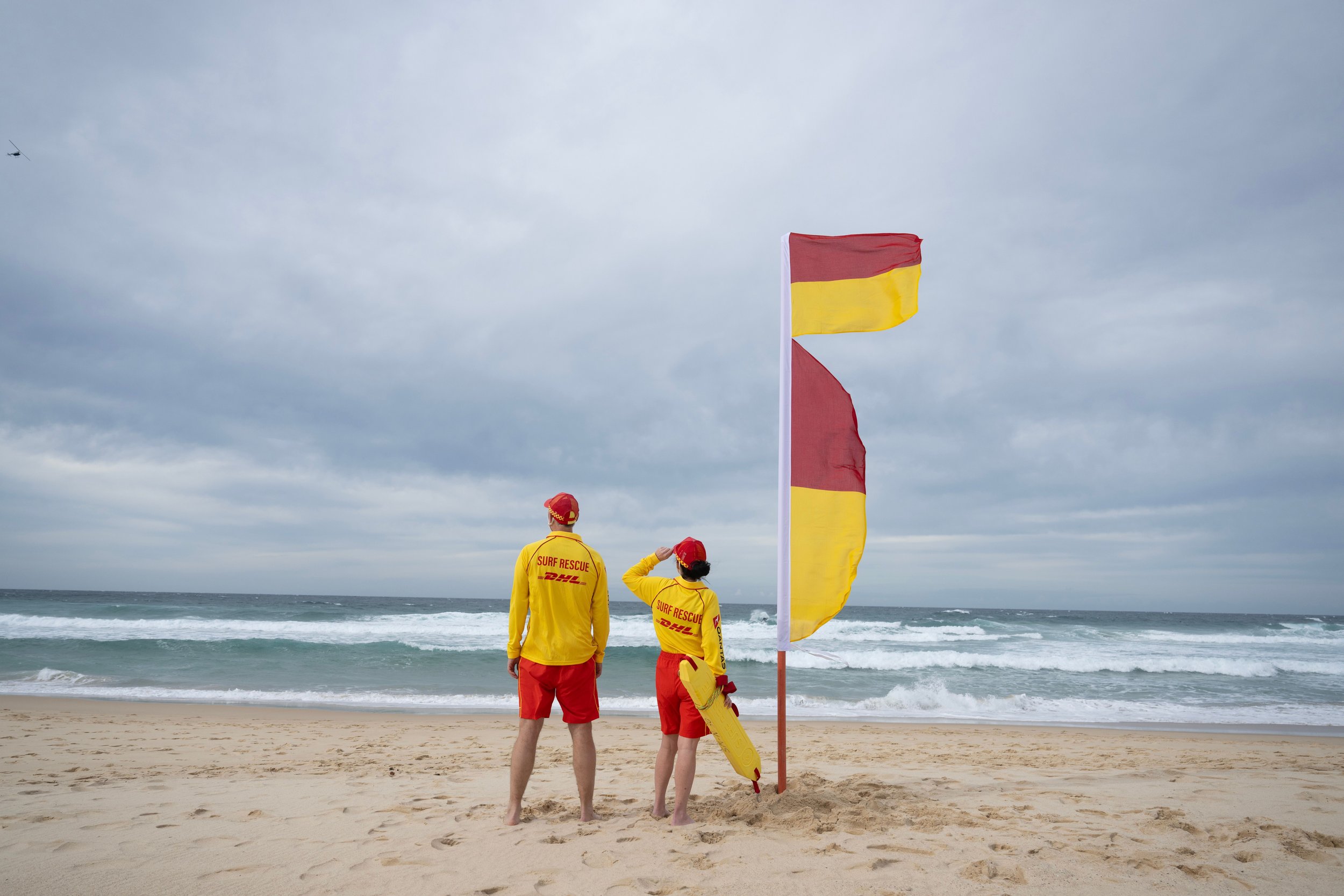 Two surf rescue team members in yellow and red uniforms standing on a sandy beach near the ocean, with one holding a yellow rescue float and another a red and yellow rescue flag, under a cloudy sky.