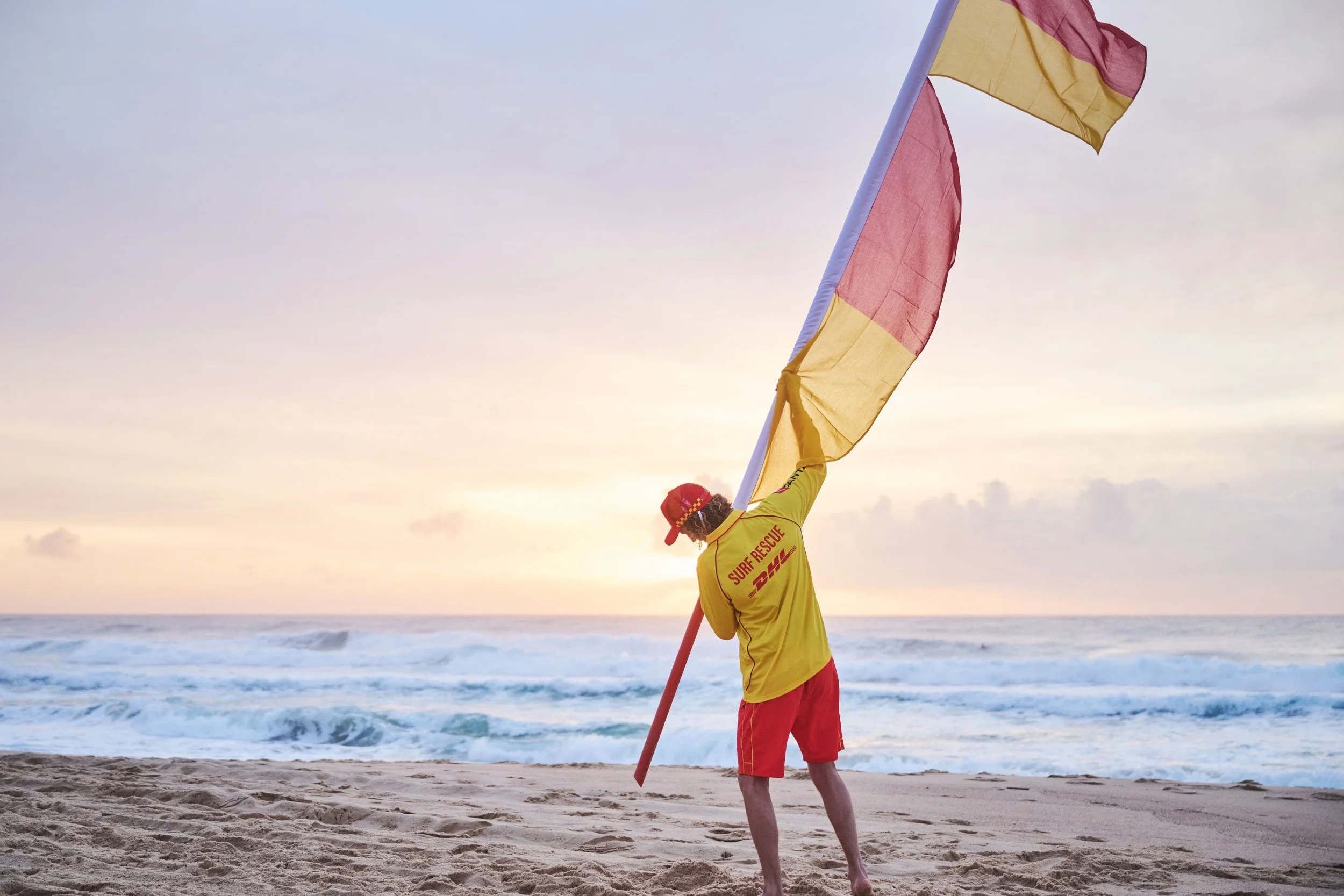 A surf rescue worker holding a yellow and red flag on a beach at sunset, with ocean waves in the background.