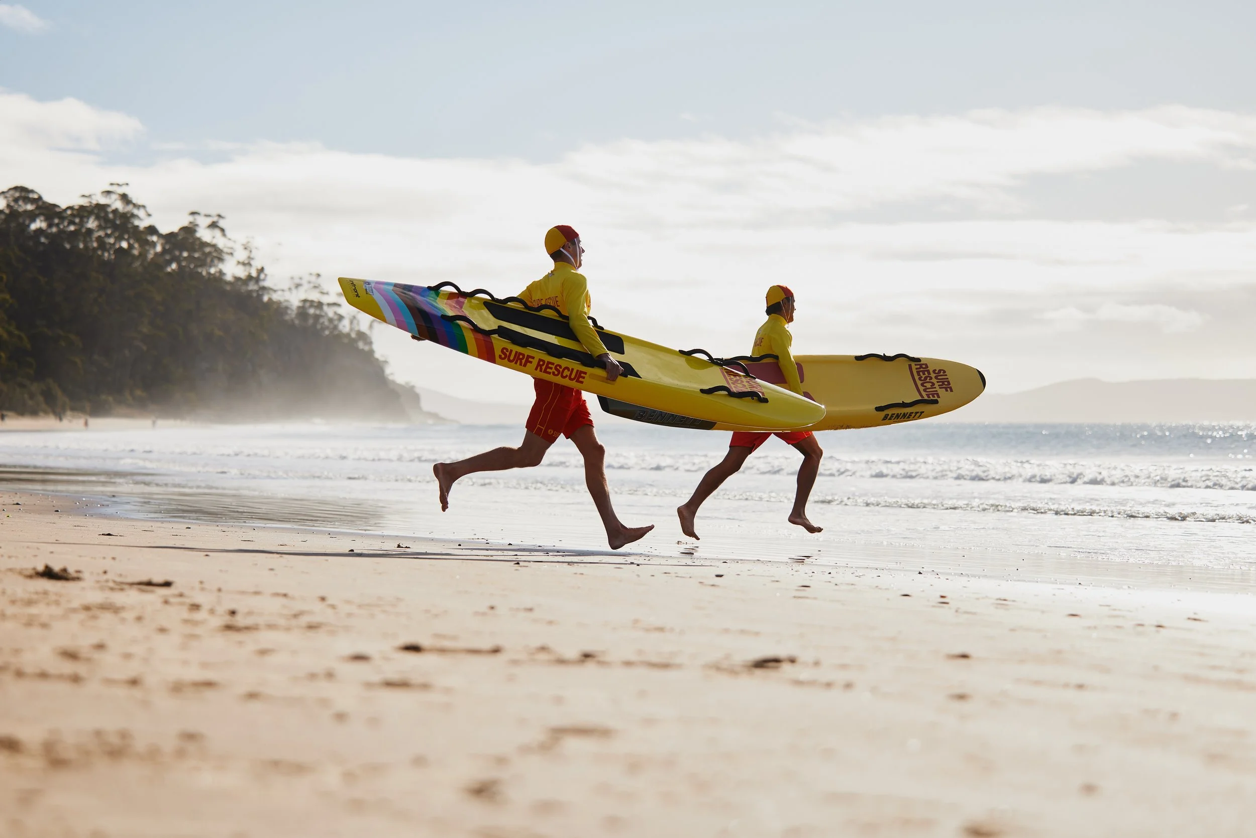 Two lifeguards in yellow shirts and red shorts running towards the water with yellow rescue boards.