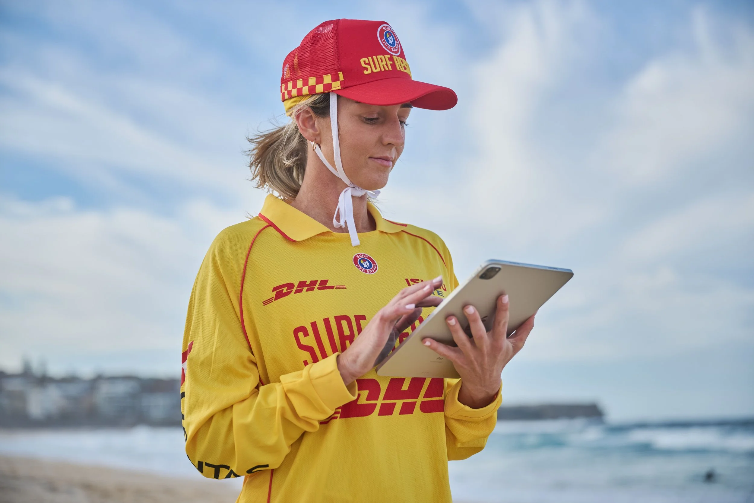 A woman in a yellow Surf Rescue uniform and red cap, holding a tablet, standing outdoors near the water under a partly cloudy sky.