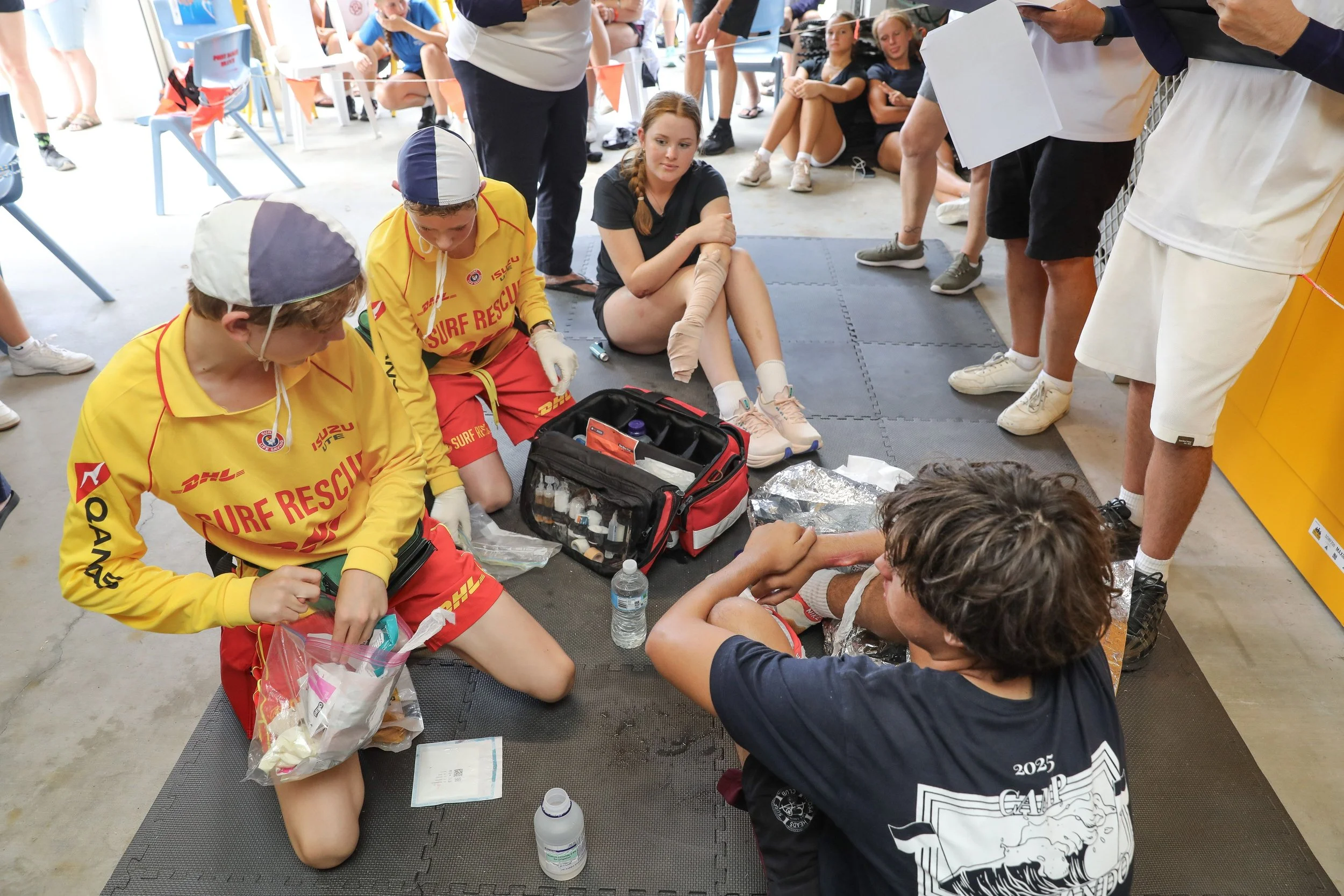 A group of young people at a first aid training session. One person is lying on the ground with a leg injury, while individuals dressed in yellow rescue uniforms tend to her, providing medical assistance. Others are gathered around, observing and taking notes.