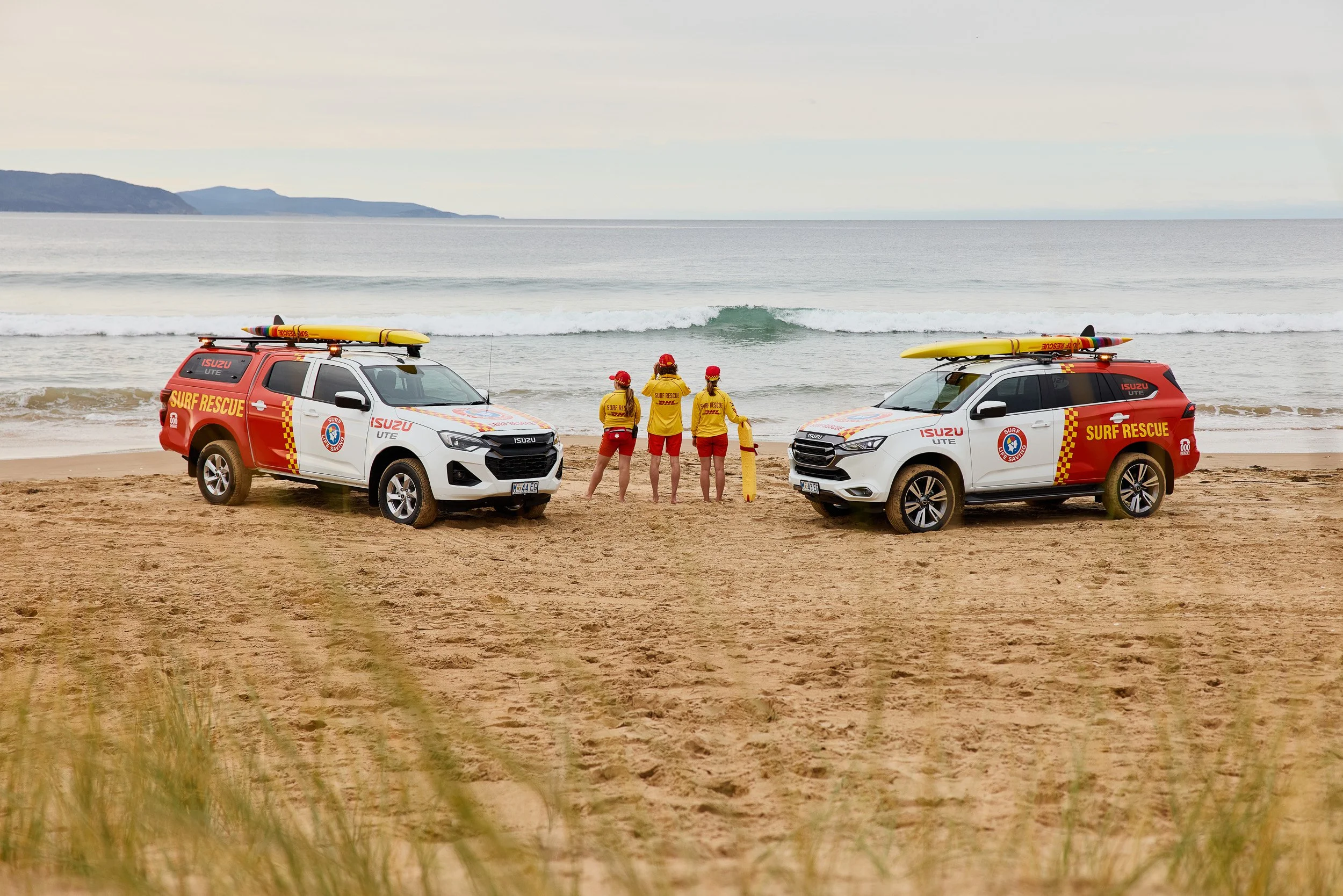 Four surf rescue team members in yellow and red uniforms standing on a sandy beach, with two emergency vehicles equipped with surfboards nearby, and the ocean with small waves in the background.