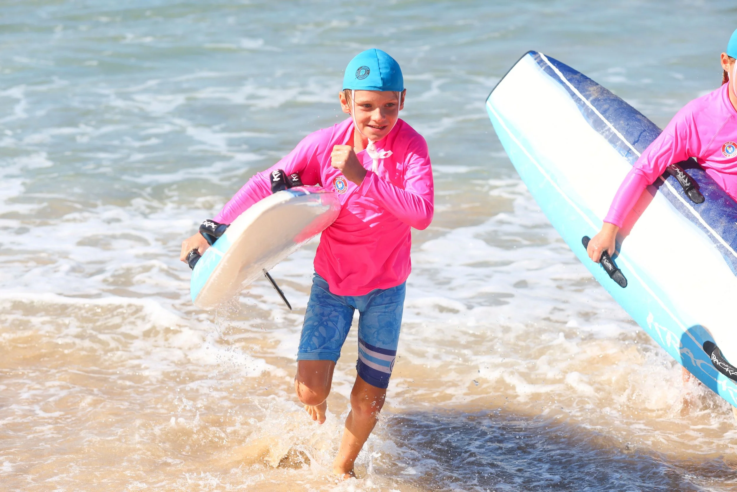 A young boy in a pink rash guard and blue shorts is running out of the water at the beach, holding a blue and white surfboard, with another person partially visible on the right holding a surfboard.