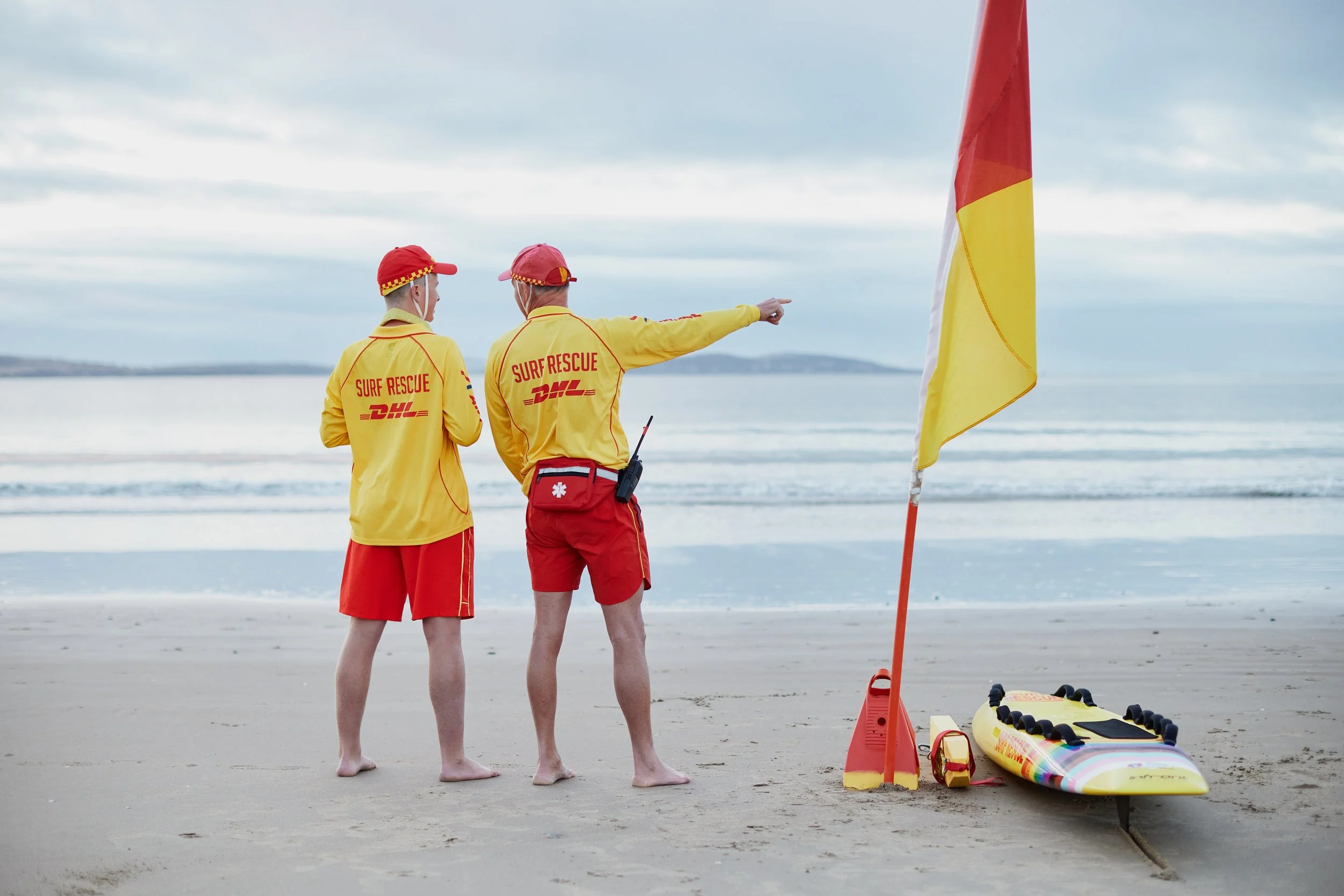 Two surf rescue team members in yellow jackets and red shorts standing on a beach, one pointing towards the ocean near a yellow and red rescue flag, with rescue equipment on the sand.