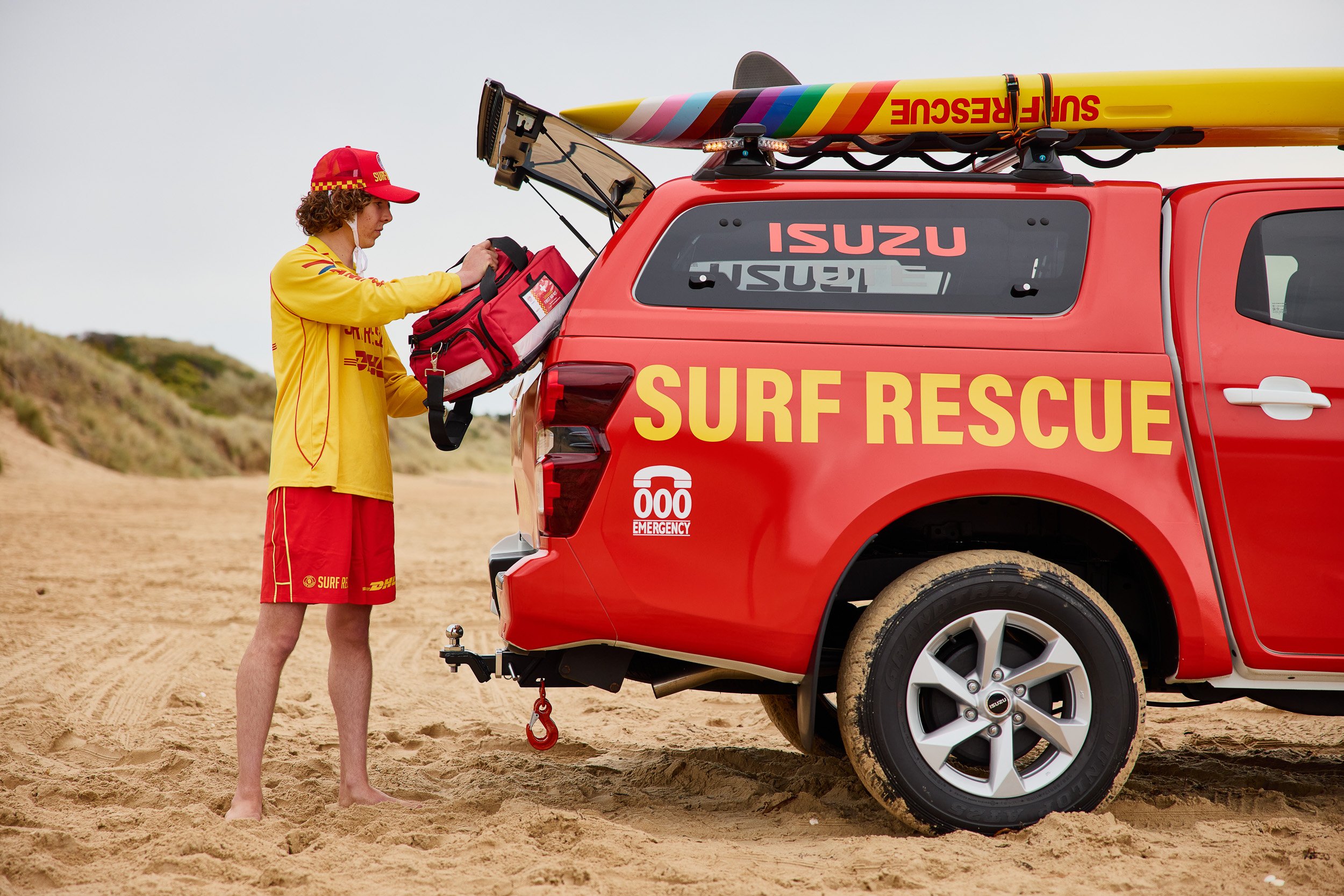 A young person in a yellow surf rescue uniform loading equipment into a red rescue vehicle on a sandy beach.