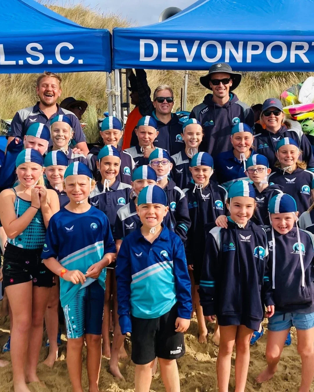 A group of young children and adults at a beach, all wearing navy blue swimwear and swim caps with a blue and white stripe, under a blue tent with the words 'L.S.C. DEVONPORT'.