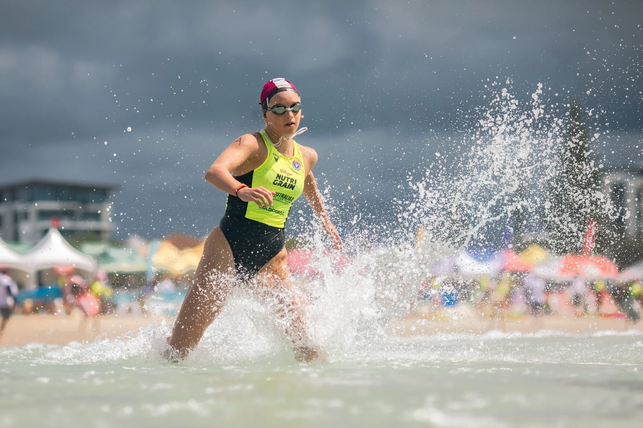 A female triathlete wearing a yellow top, black swim cap, and goggles running out of the water during a race, with splashes around her and a cloudy sky in the background.