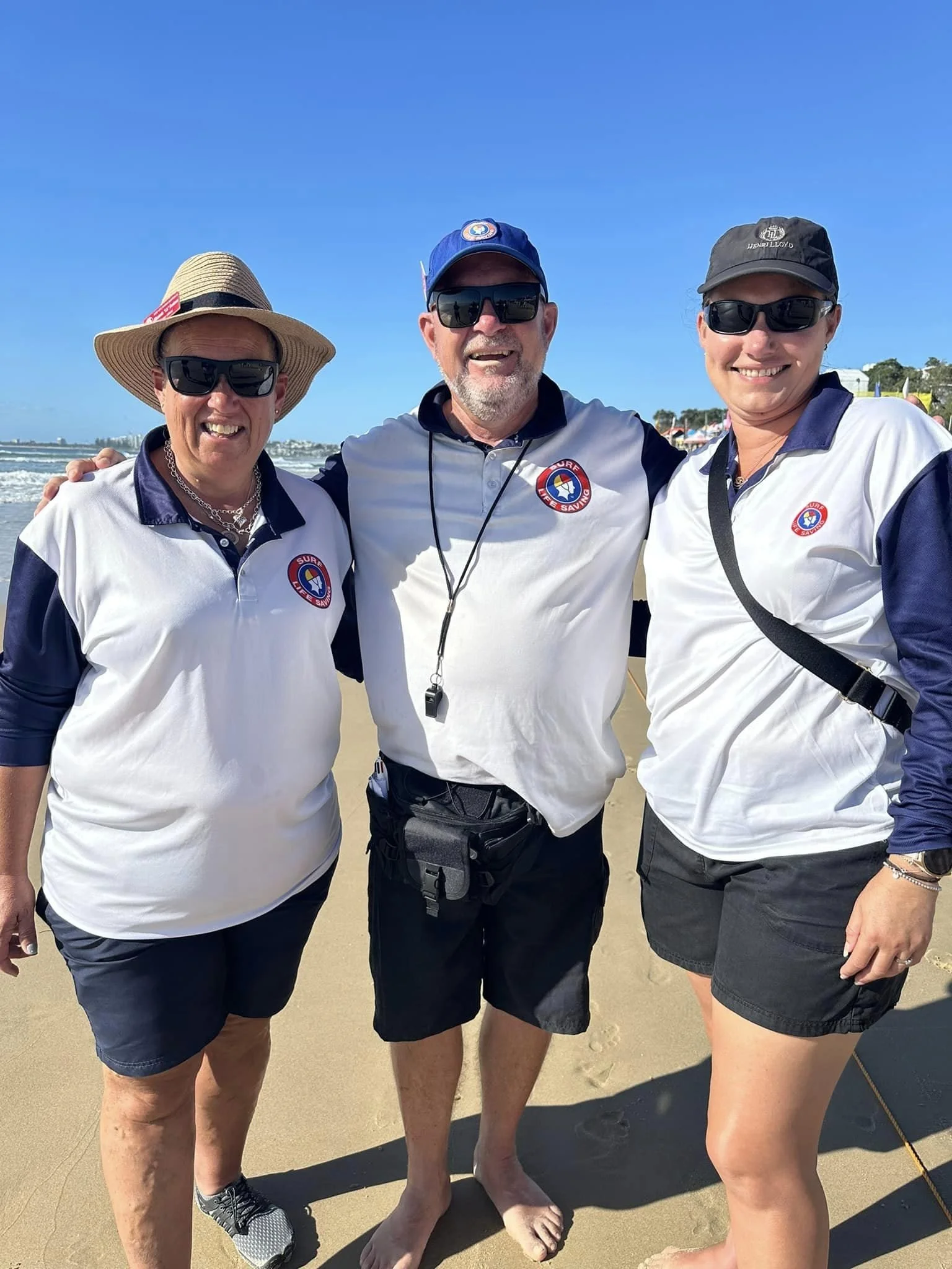 Three people standing together at the beach, smiling, wearing matching surf life saving shirts, sunglasses, and hats, with the ocean and beach in the background.