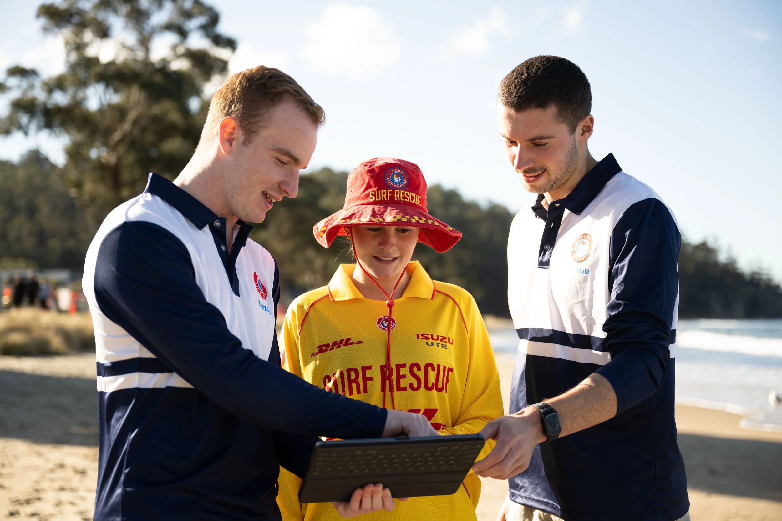 Two men and a young girl in a yellow shirt and red hat with 'Surf Rescue' written on it, looking at a tablet outdoors on a sunny day near water, with trees in the background.