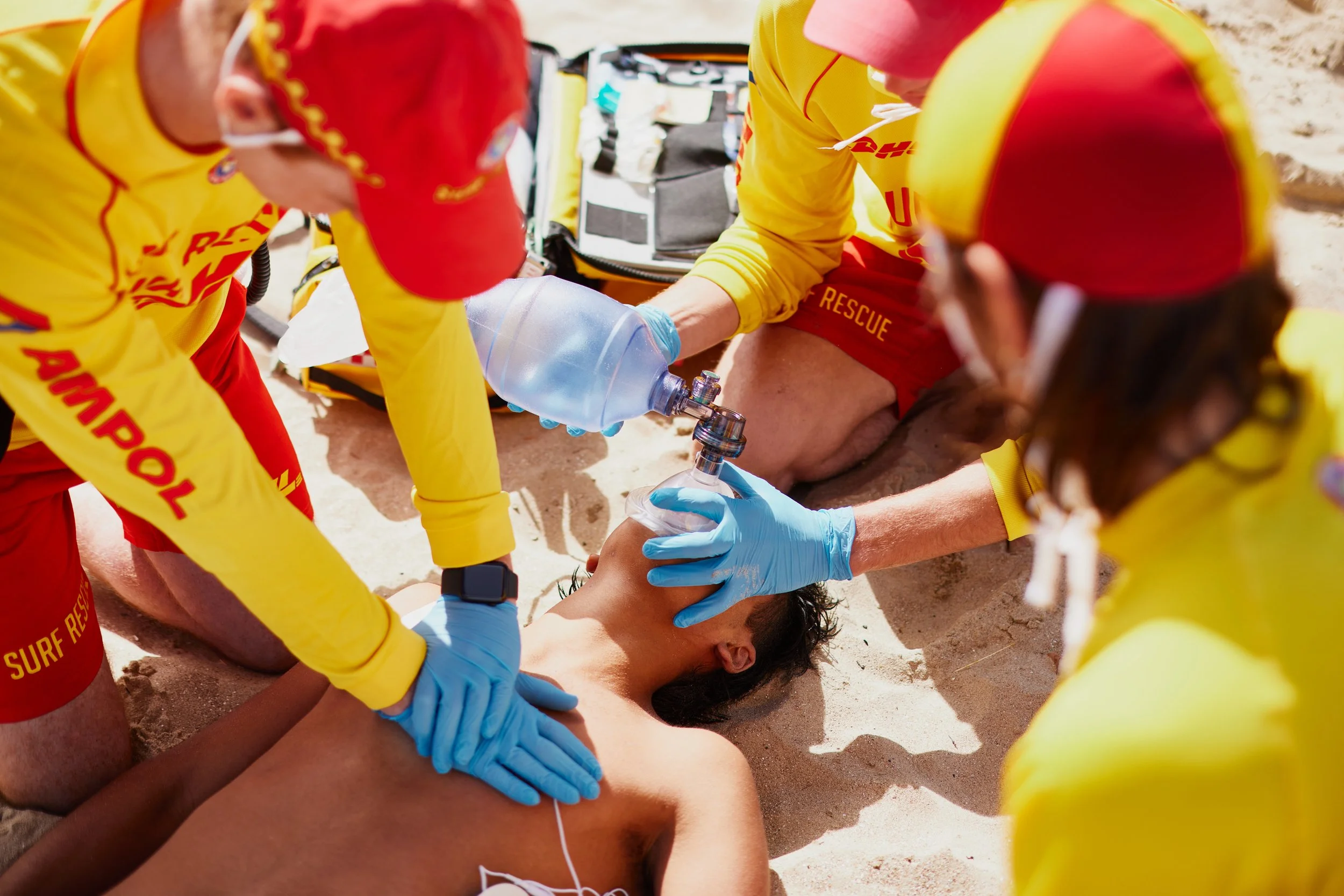 Rescue team members in yellow and red uniforms providing CPR to an unconscious person on a beach, with medical equipment nearby.