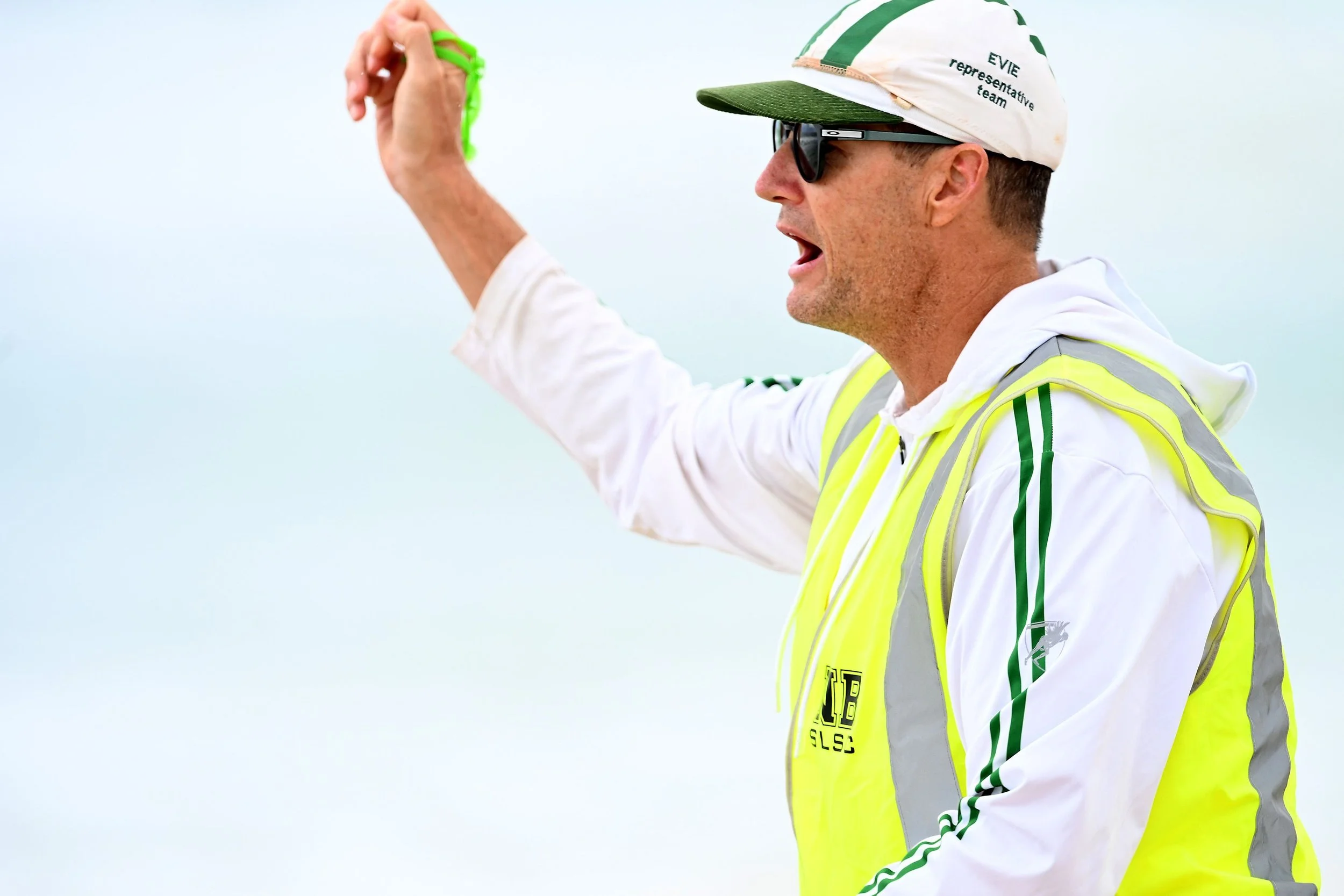 A man wearing sunglasses, a white cap with green accents and text, a white hoodie, and a yellow safety vest is holding a green object in his raised right hand, with a light blue sky in the background.