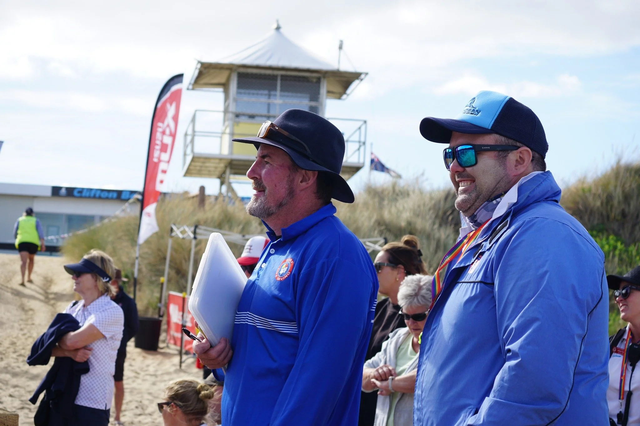 Group of people standing outdoors near a beach with a lifeguard tower and flags, wearing casual and sports attire, some with sunglasses and hats, smiling and observing an event.