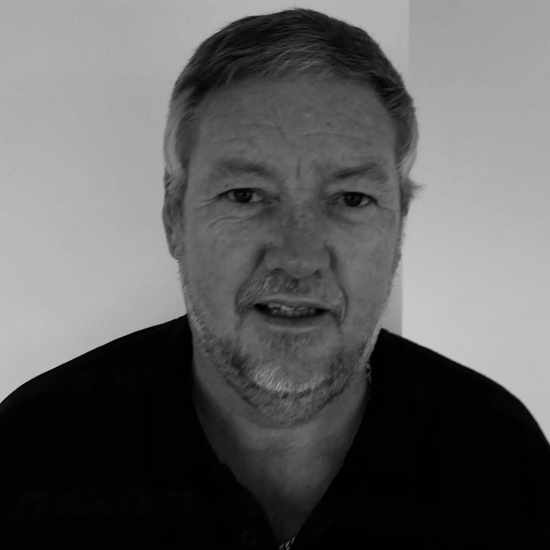 Close-up black and white photo of an elderly man with short, grey hair and a beard, wearing a dark shirt.