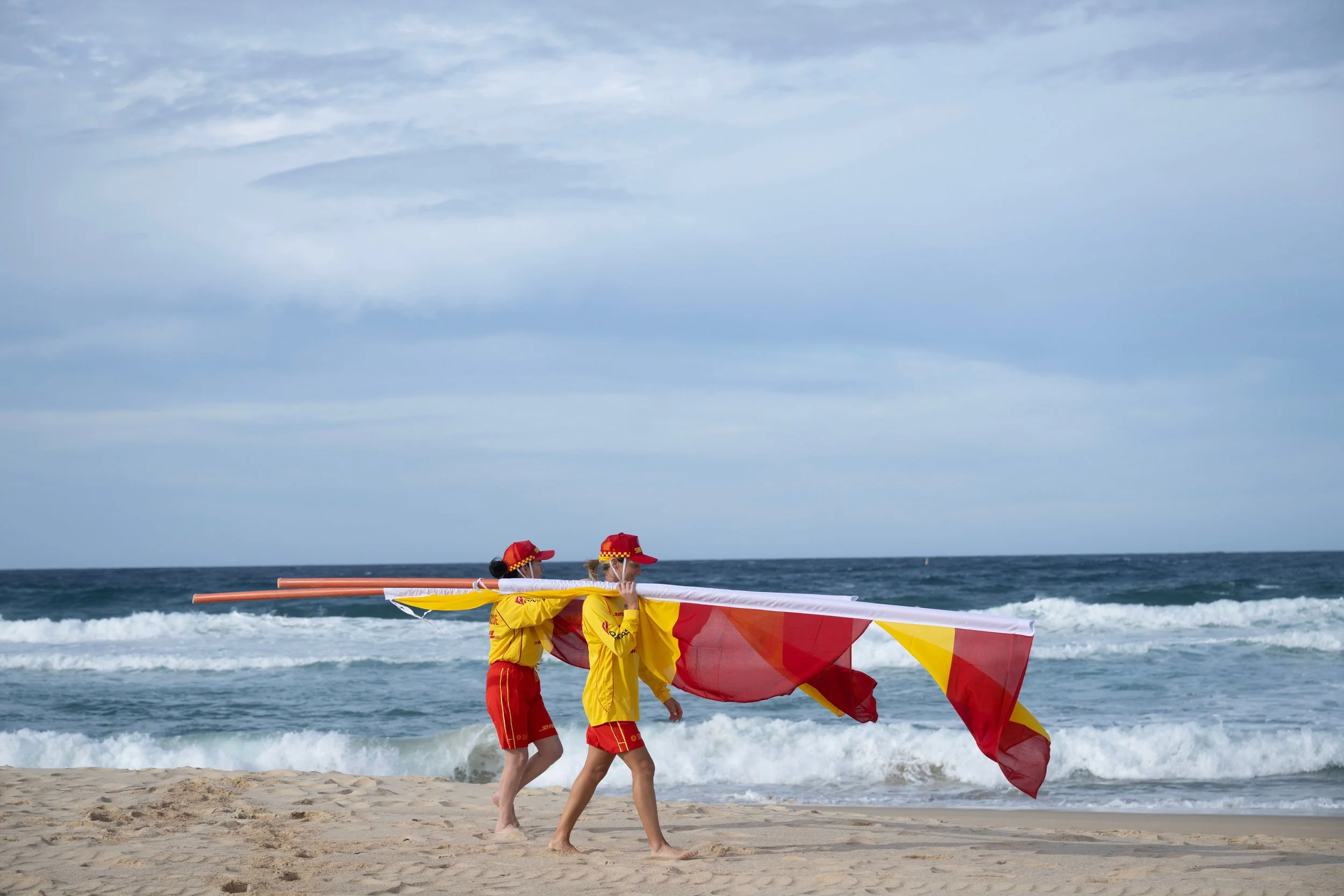 Two individuals dressed in yellow and red uniforms are on a beach, carrying a large red and yellow flag. They are near the ocean with waves and a cloudy sky in the background.